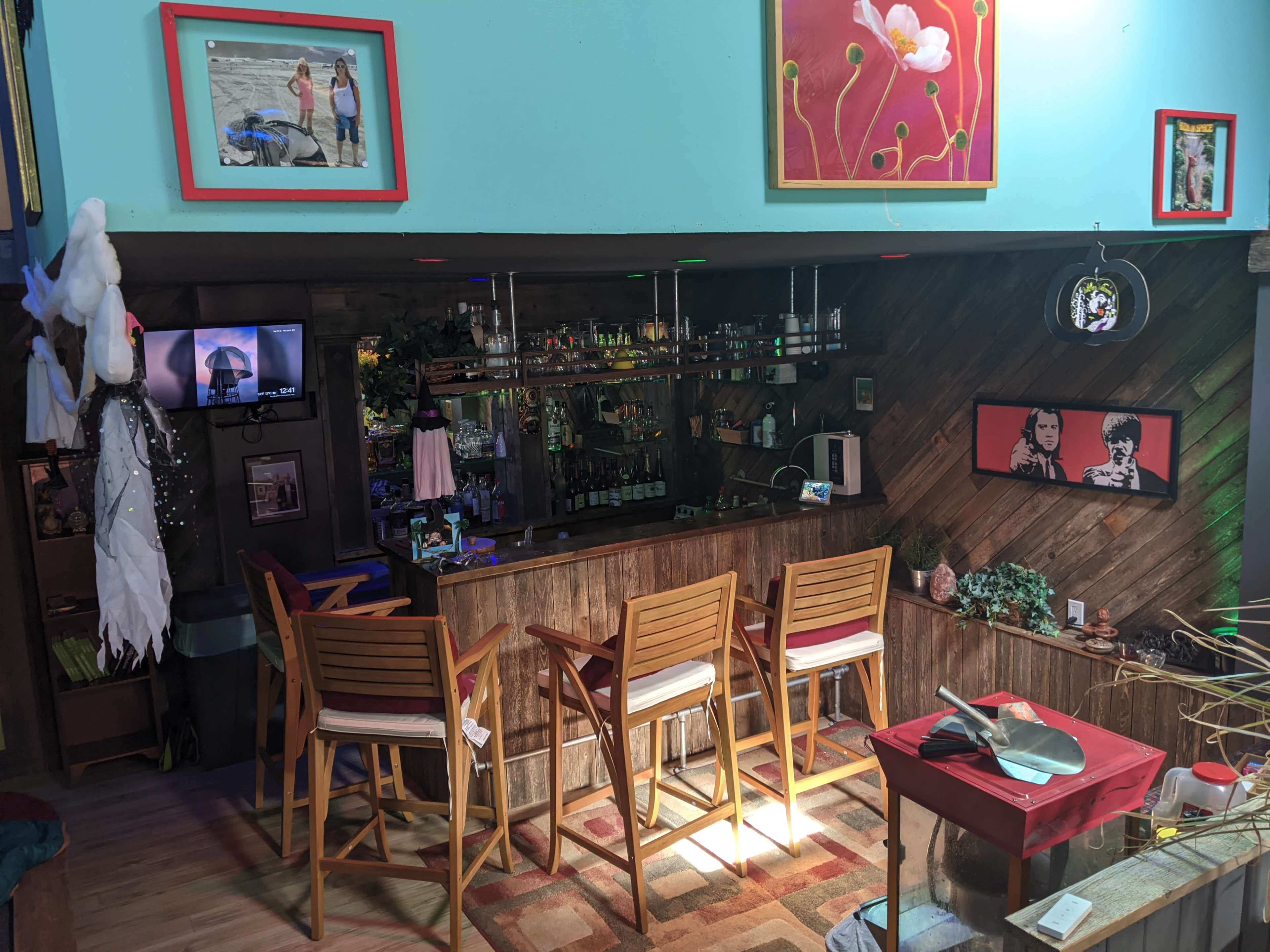 A cozy bar area with wooden stools, a variety of bottles on shelves, and colorful artwork hanging on the walls.