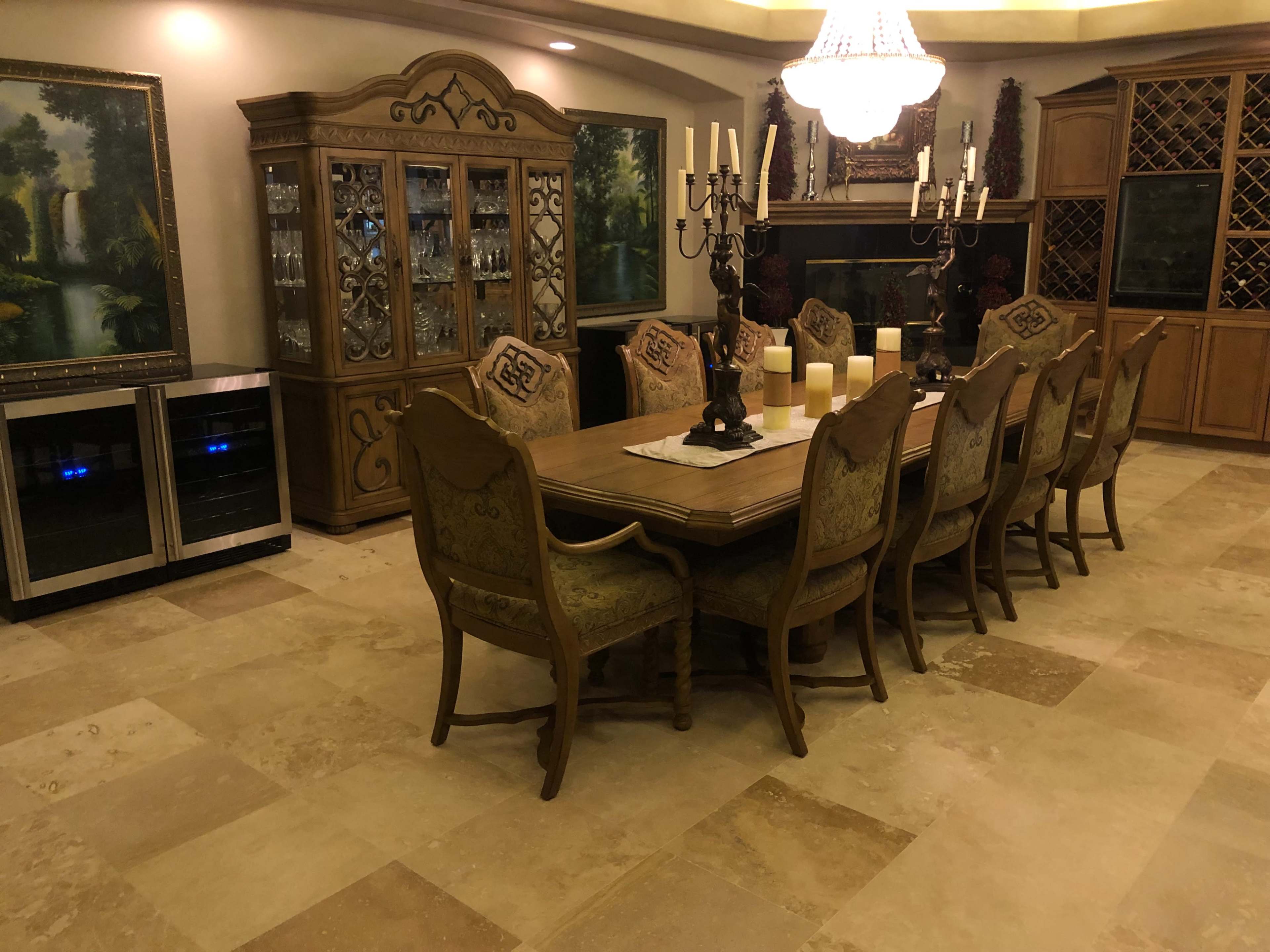 The image shows a dining room with a large wooden table surrounded by eight upholstered chairs, a china cabinet, and a wine cabinet, with a chandelier hanging overhead.