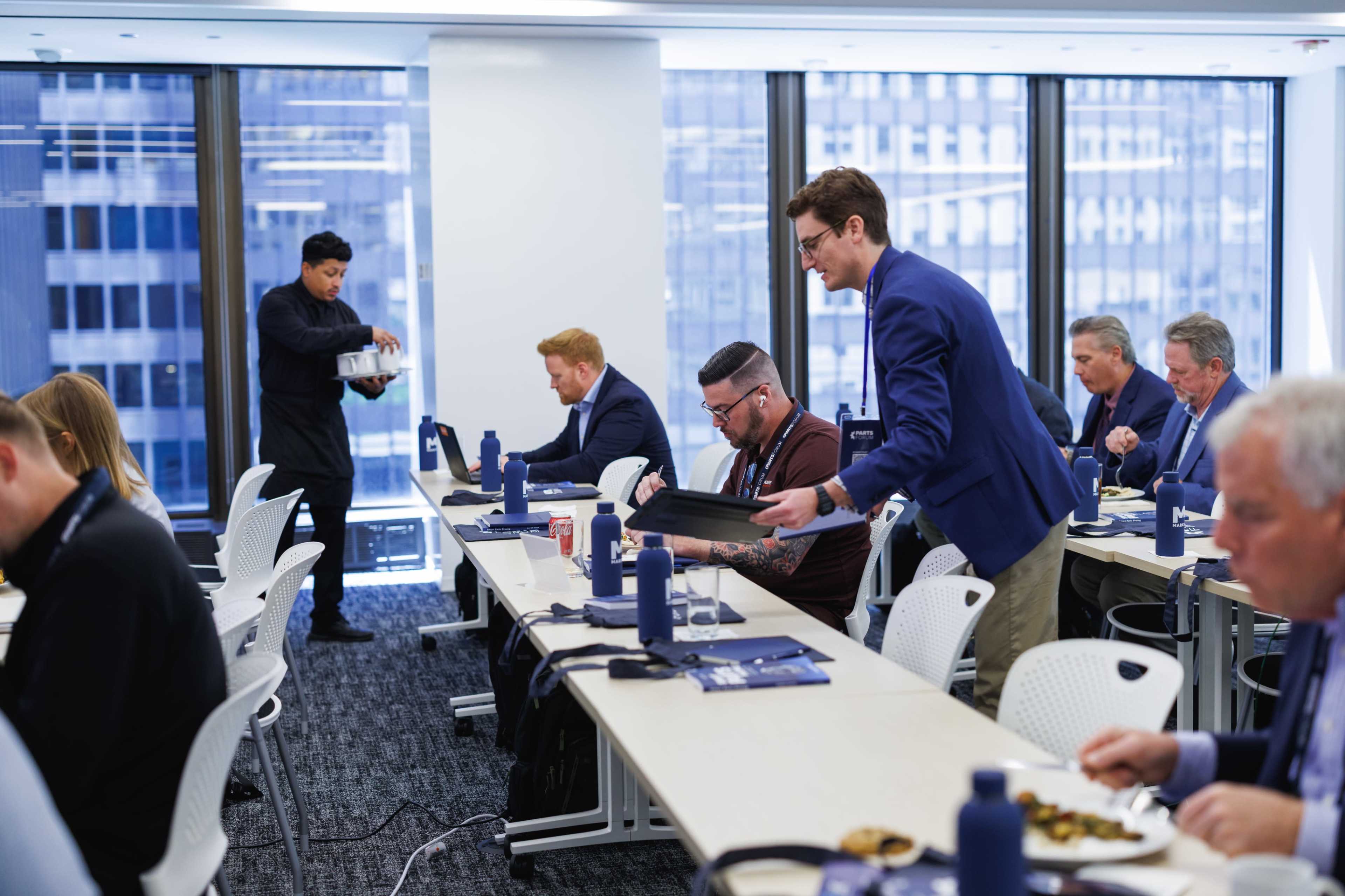 A group of professionals sits at long tables in a conference room while one person serves food to a seated attendee.