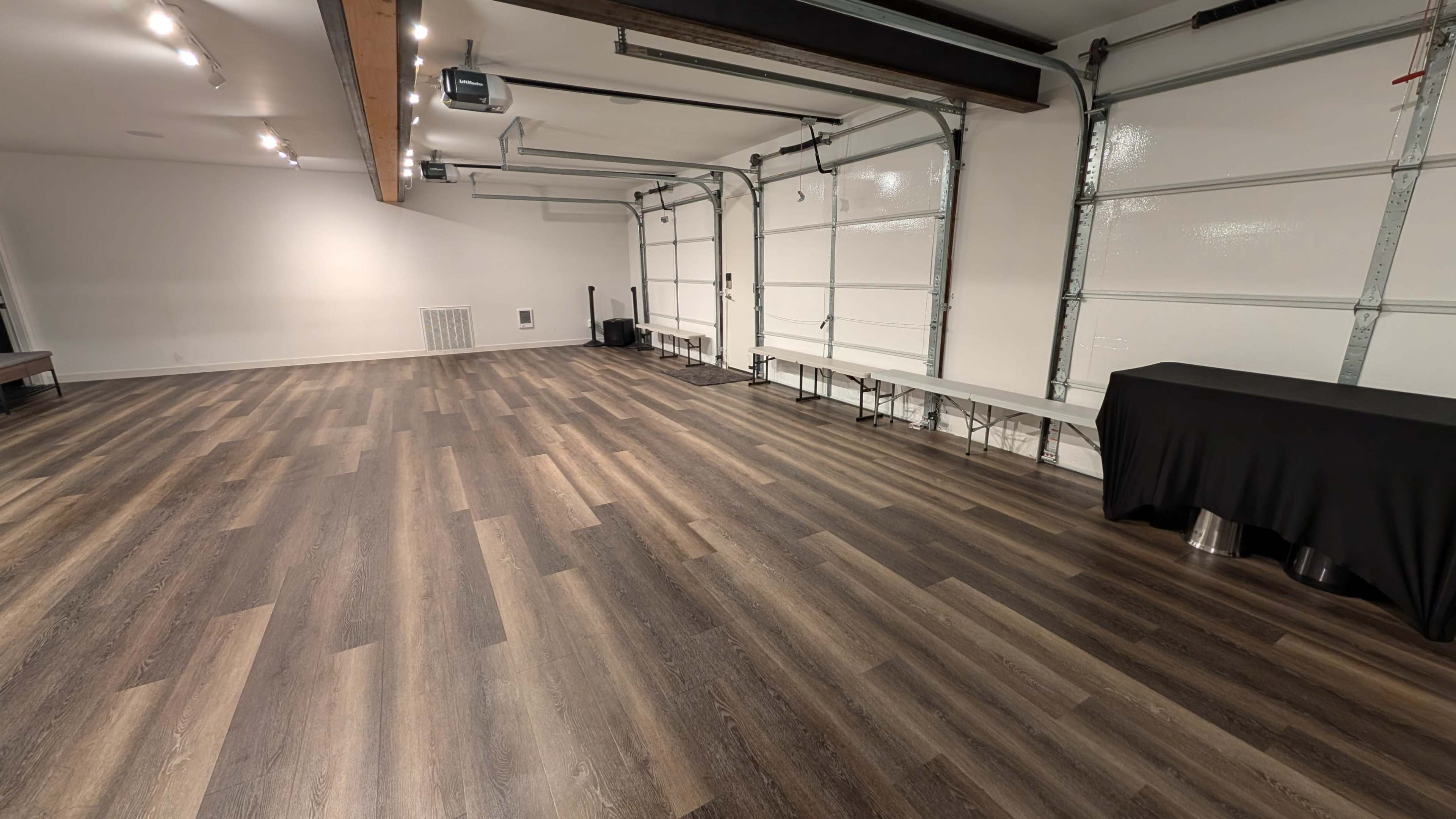 The image shows a spacious interior with a polished wooden floor, white walls, and a garage-style door, featuring minimal furniture along one side.