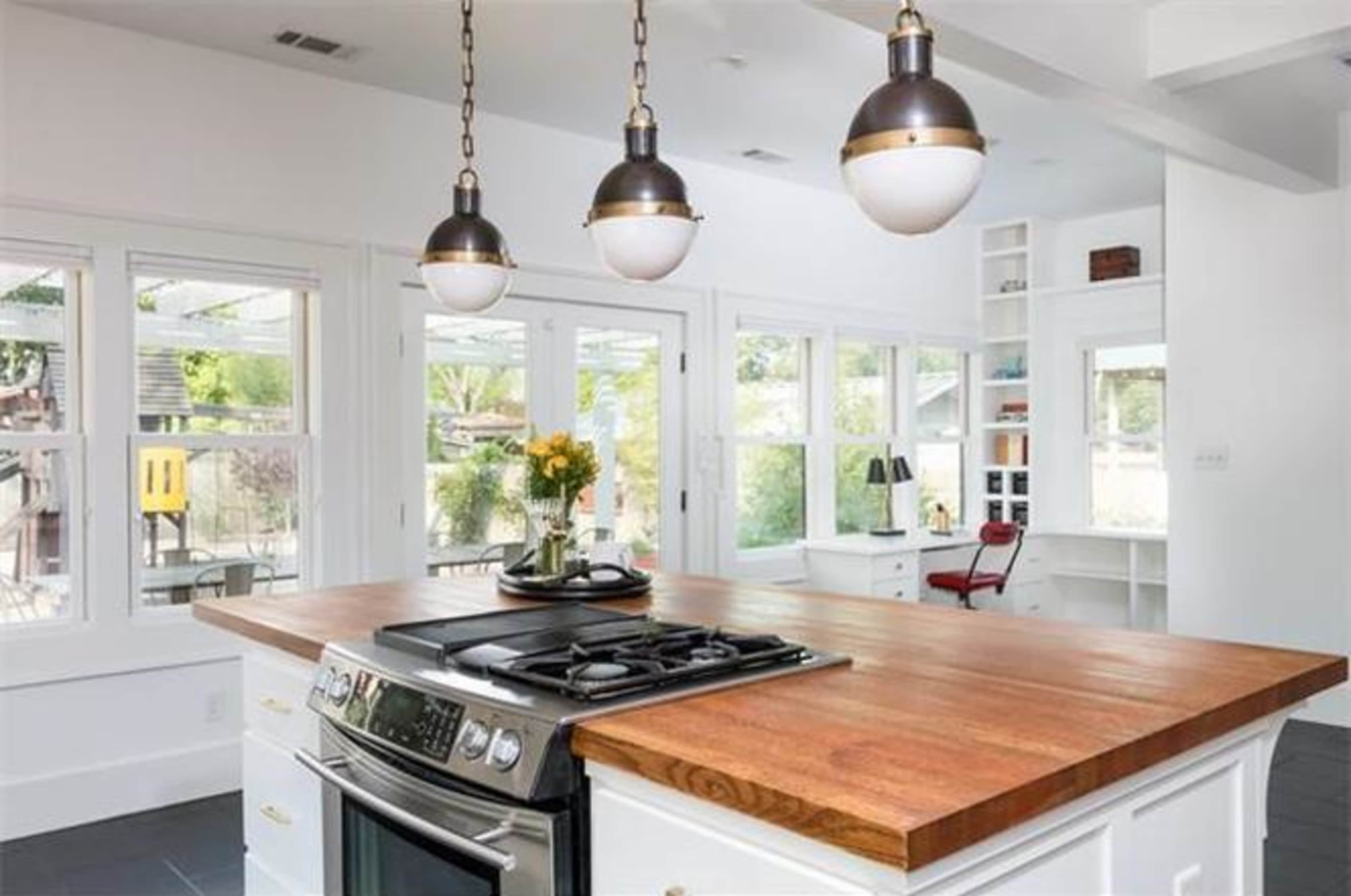 A modern kitchen features a wooden island with a stove, three pendant lights overhead, and large windows offering a view of the outdoor area.