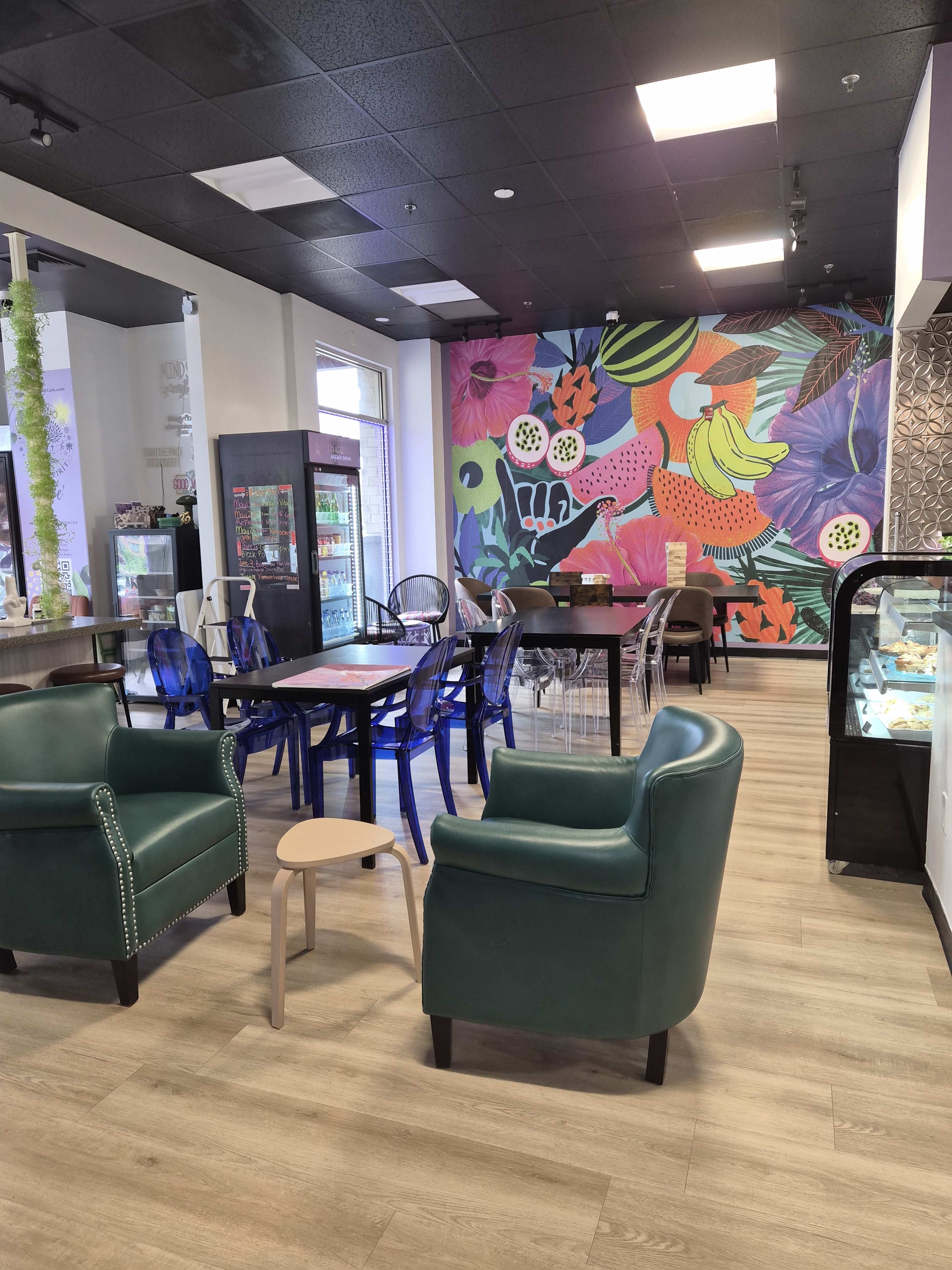 The image shows a modern café with colorful floral artwork on the back wall, comfortable seating areas, and a display case for pastries.