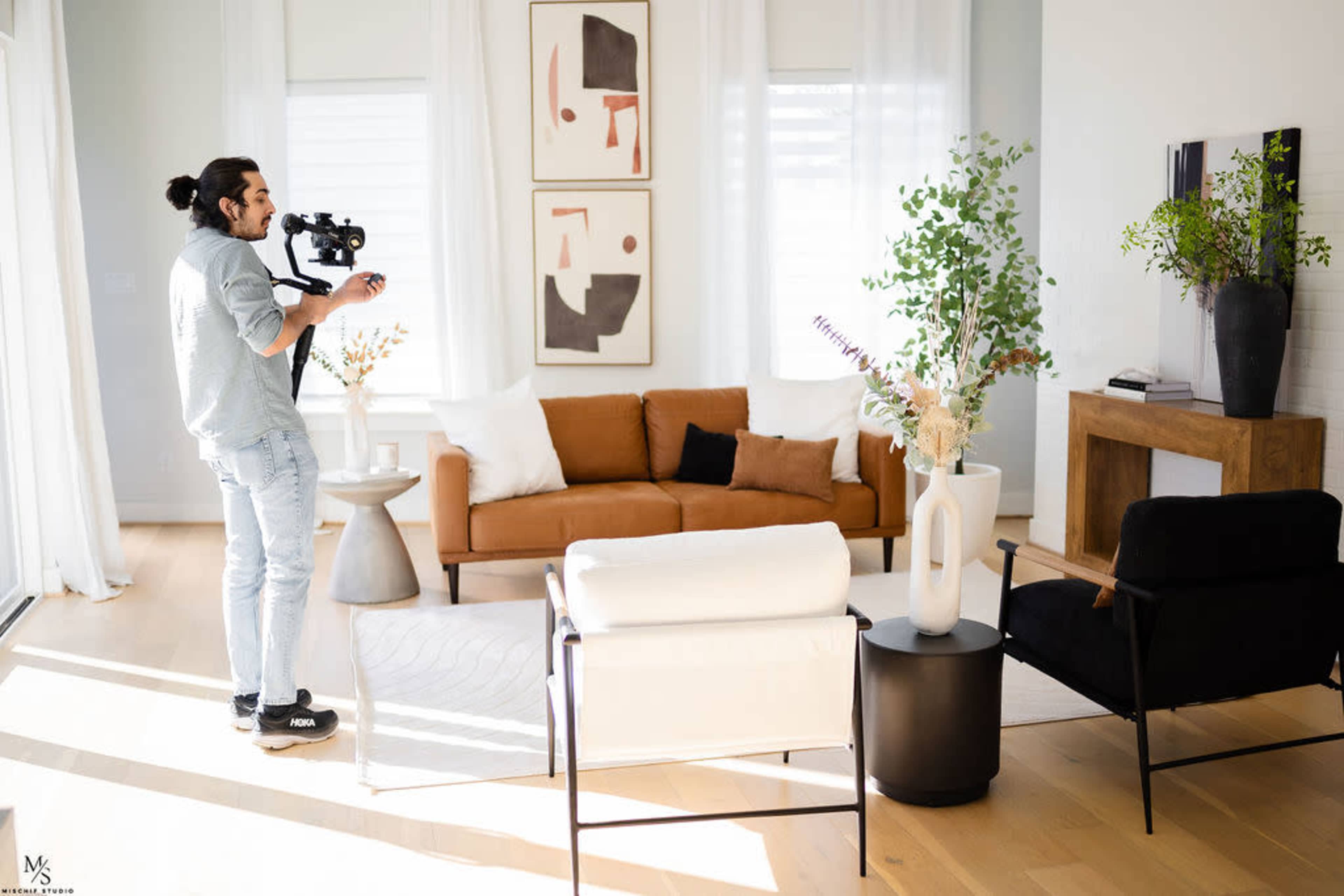 A man holds a video camera while standing in a brightly lit living room with a brown sofa, a white chair, and decorative plants.