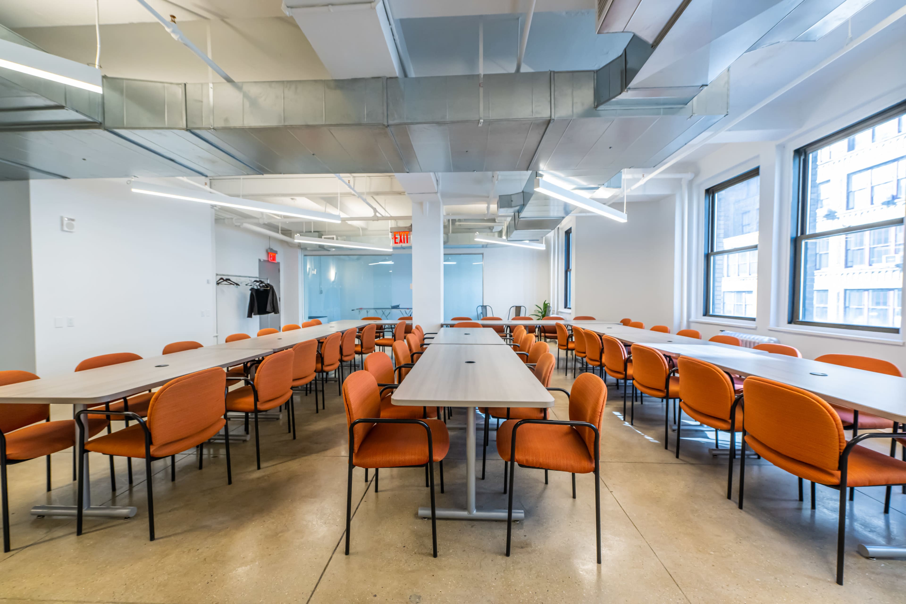 A modern meeting room with multiple long tables surrounded by orange chairs, illuminated by overhead lighting and featuring large windows.