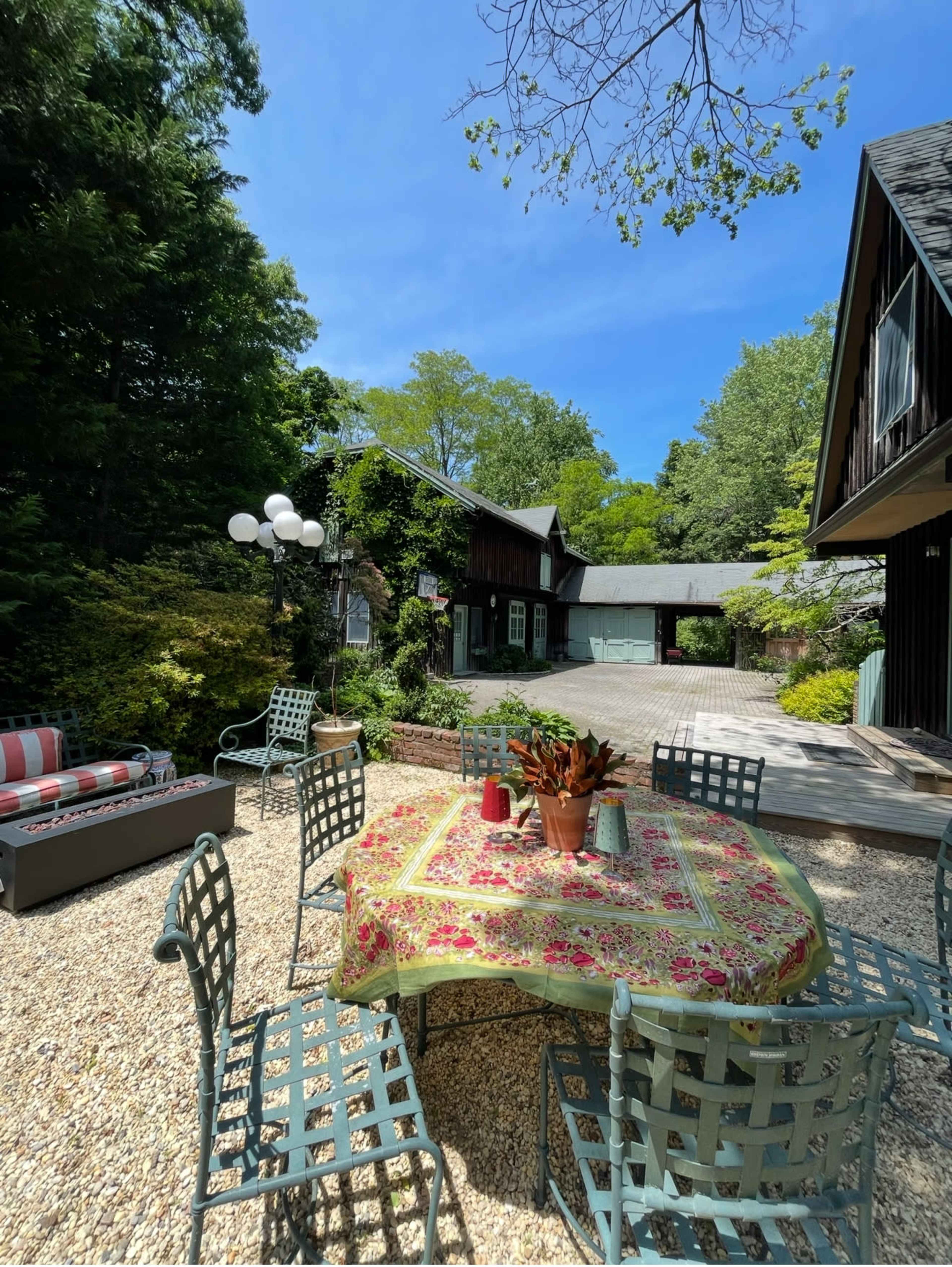A patio area featuring a table set with a floral tablecloth and surrounded by green metal chairs, adjacent to a house and a garage under a clear blue sky.