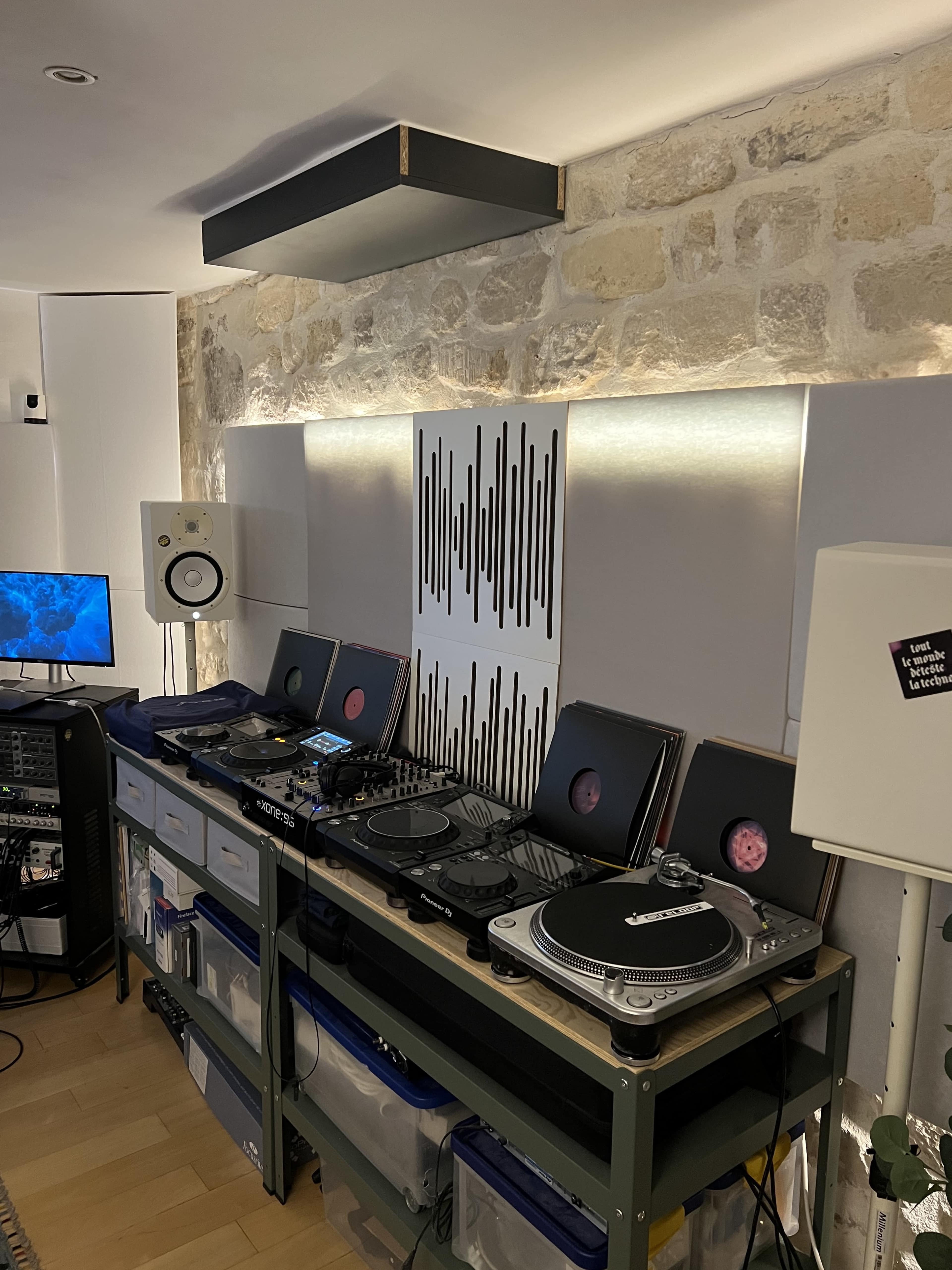 The image shows a music production or DJ setup with turntables, a mixer, speakers, and vinyl records arranged on a shelving unit against a textured wall.