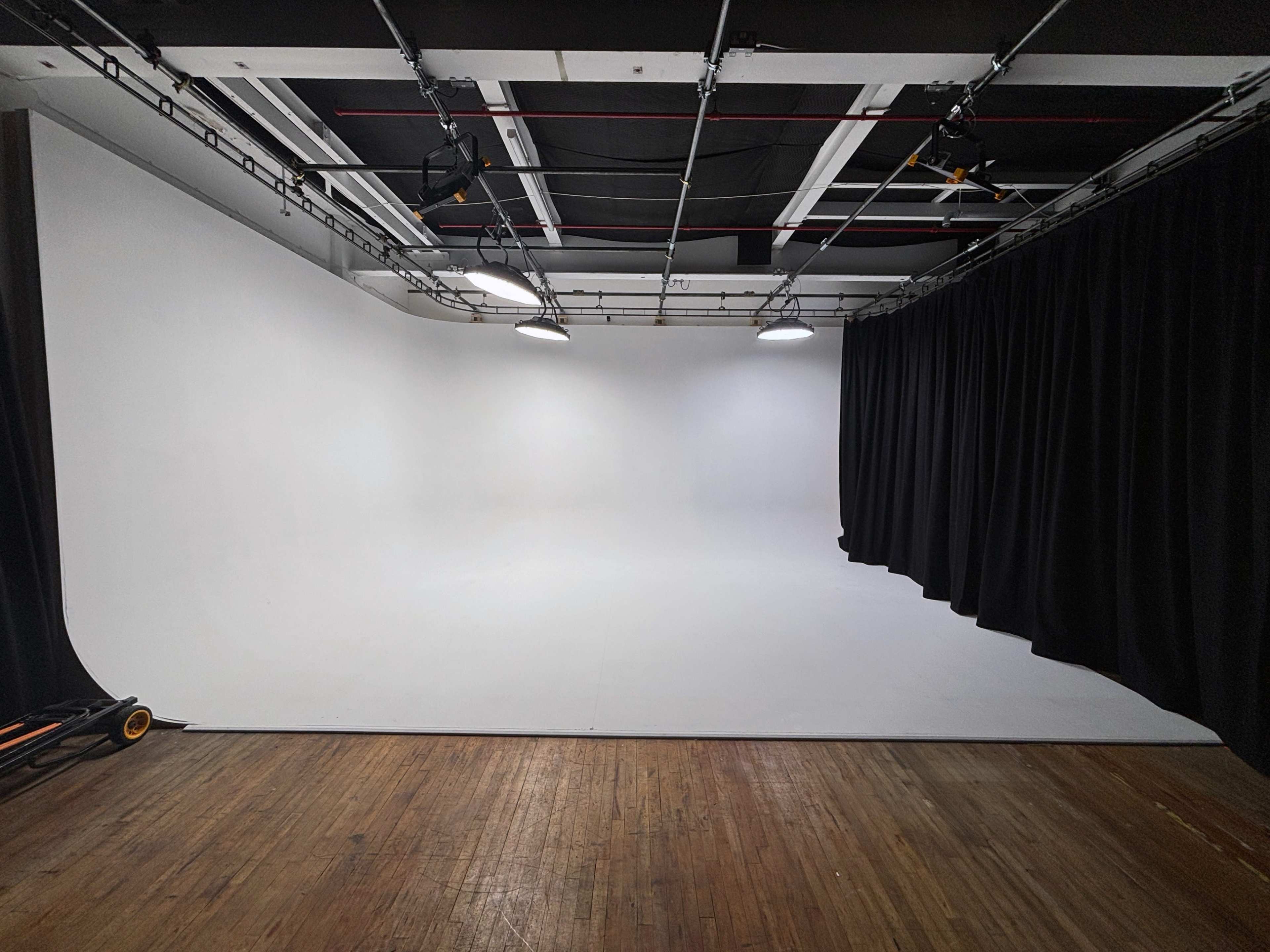 The image shows a photo studio with a white backdrop and black curtains, illuminated by overhead lights, and featuring a wooden floor.