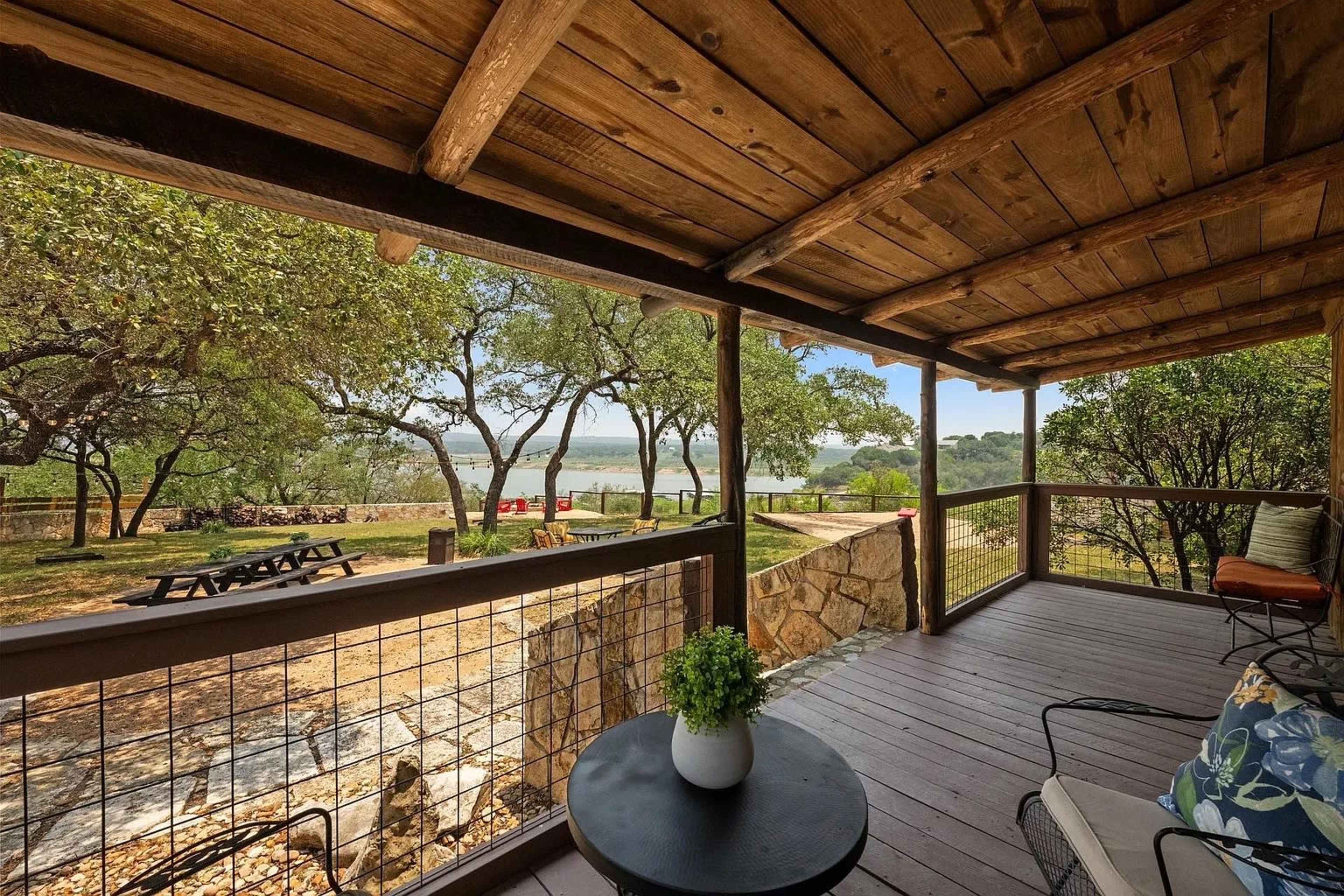 A wooden porch with a table and chairs overlooks a grassy area and a body of water framed by trees.