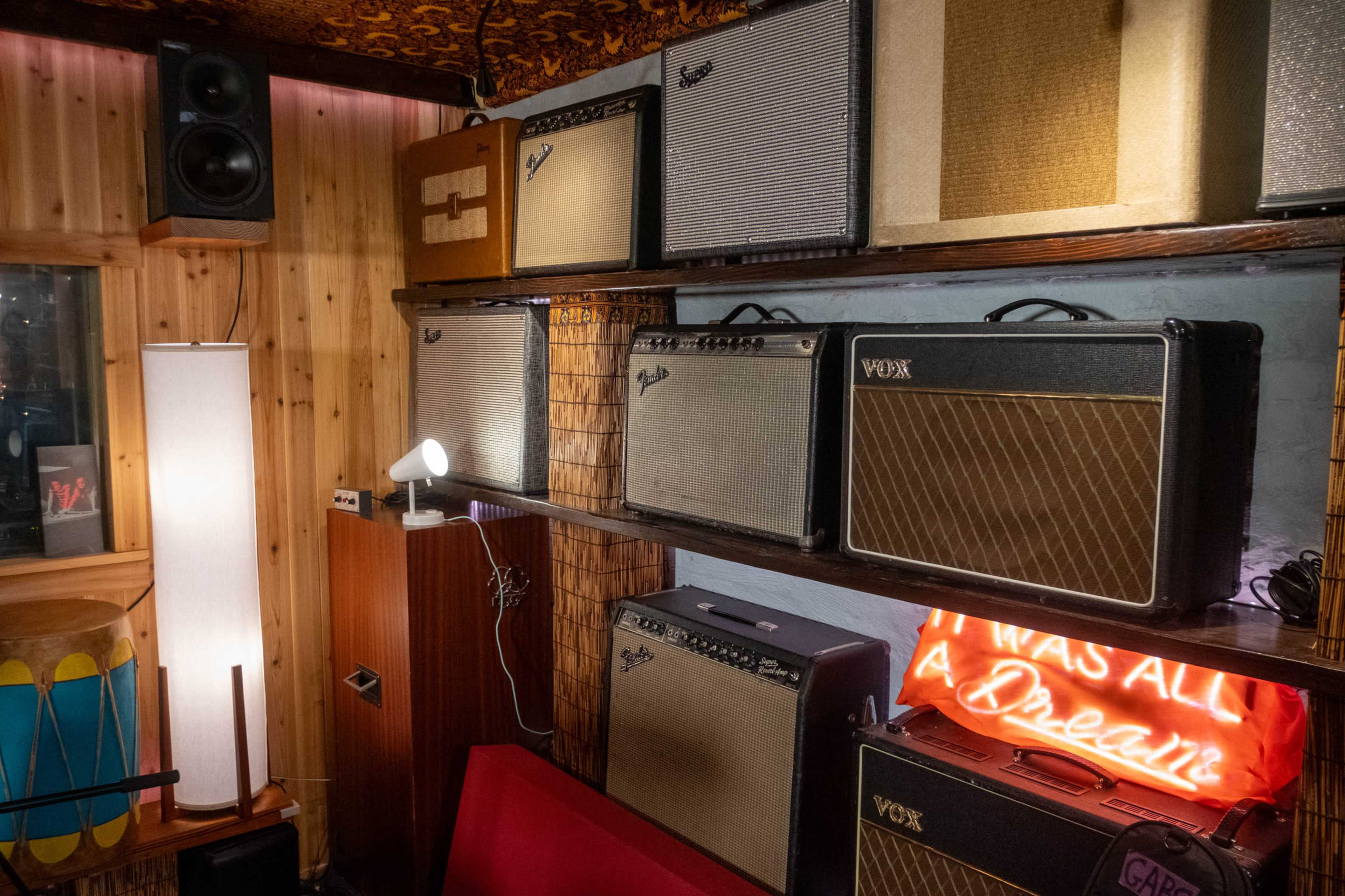 The image shows a wall mounted with various vintage amplifiers, accompanied by a glowing neon sign that reads "I WAS ALL A DREAM."