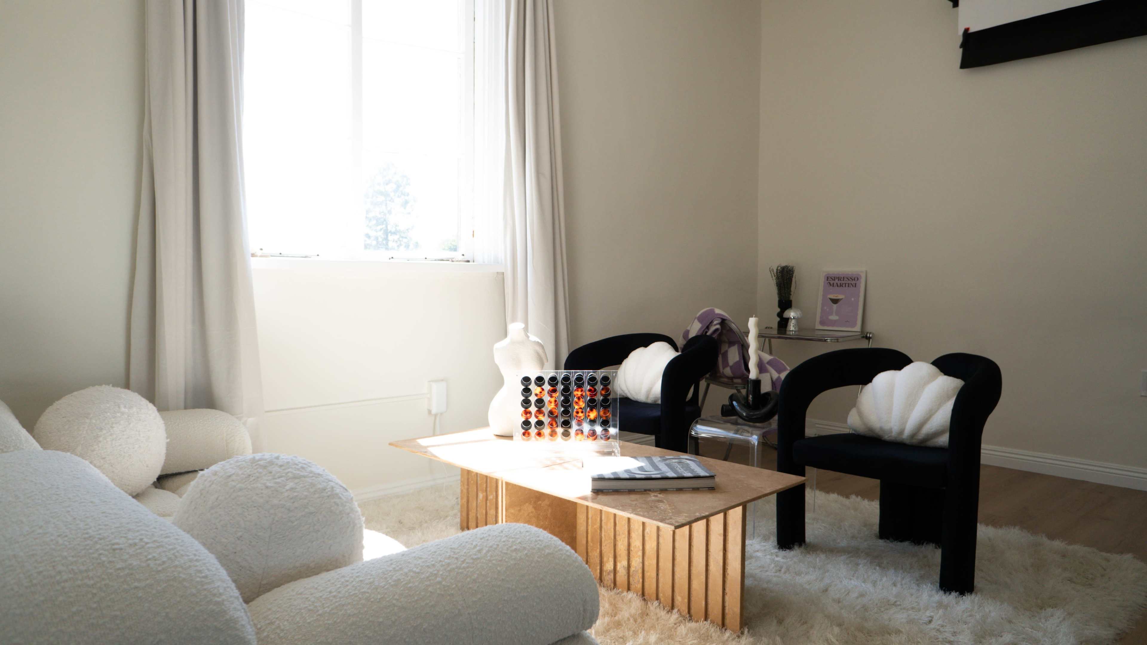 The scene features a bright room with a plush white area rug, a wooden coffee table, two black chairs, and a window that lets in natural light.