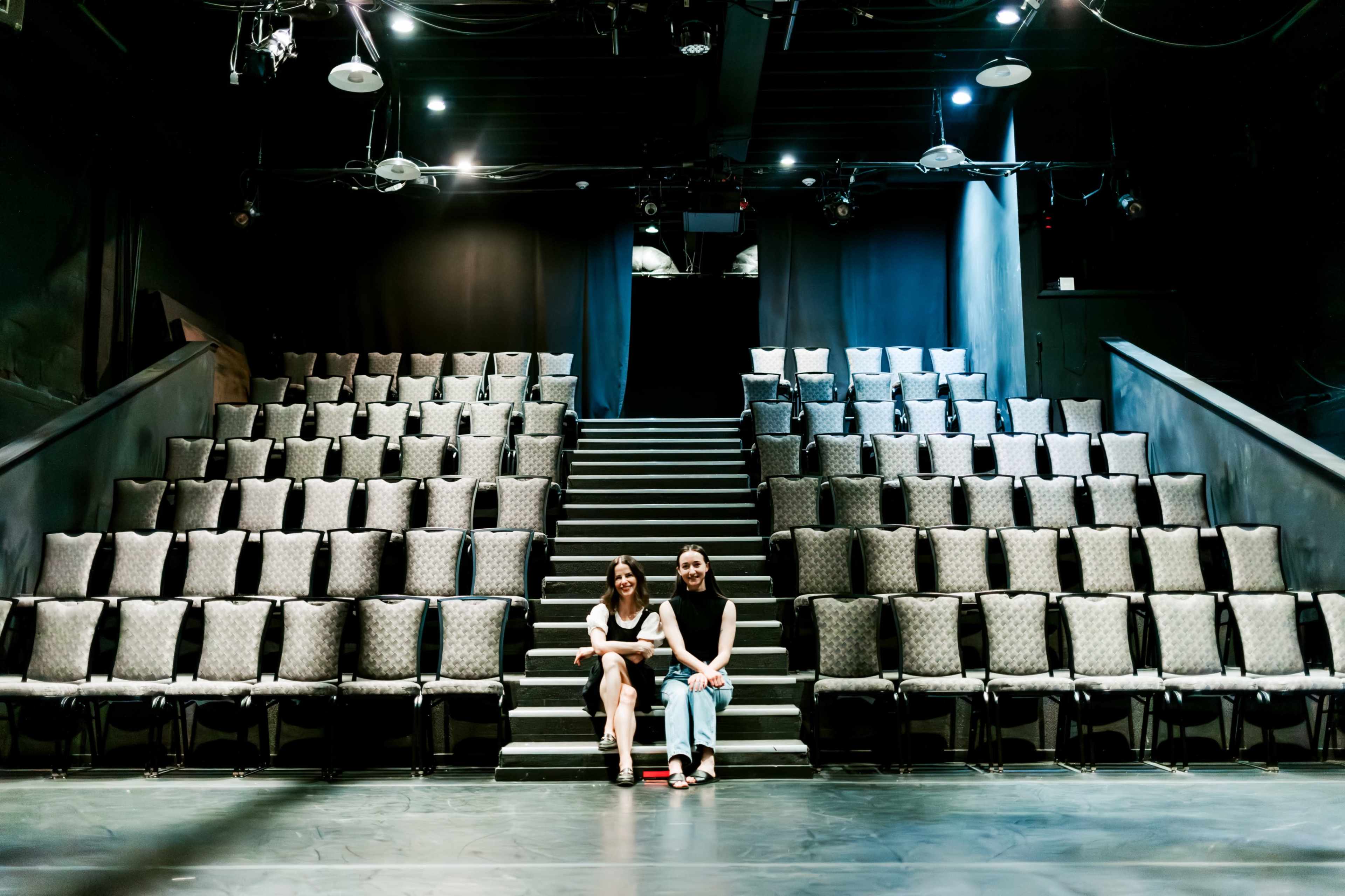 Two women sit on the stage of a theater facing an empty audience seating area filled with rows of chairs.