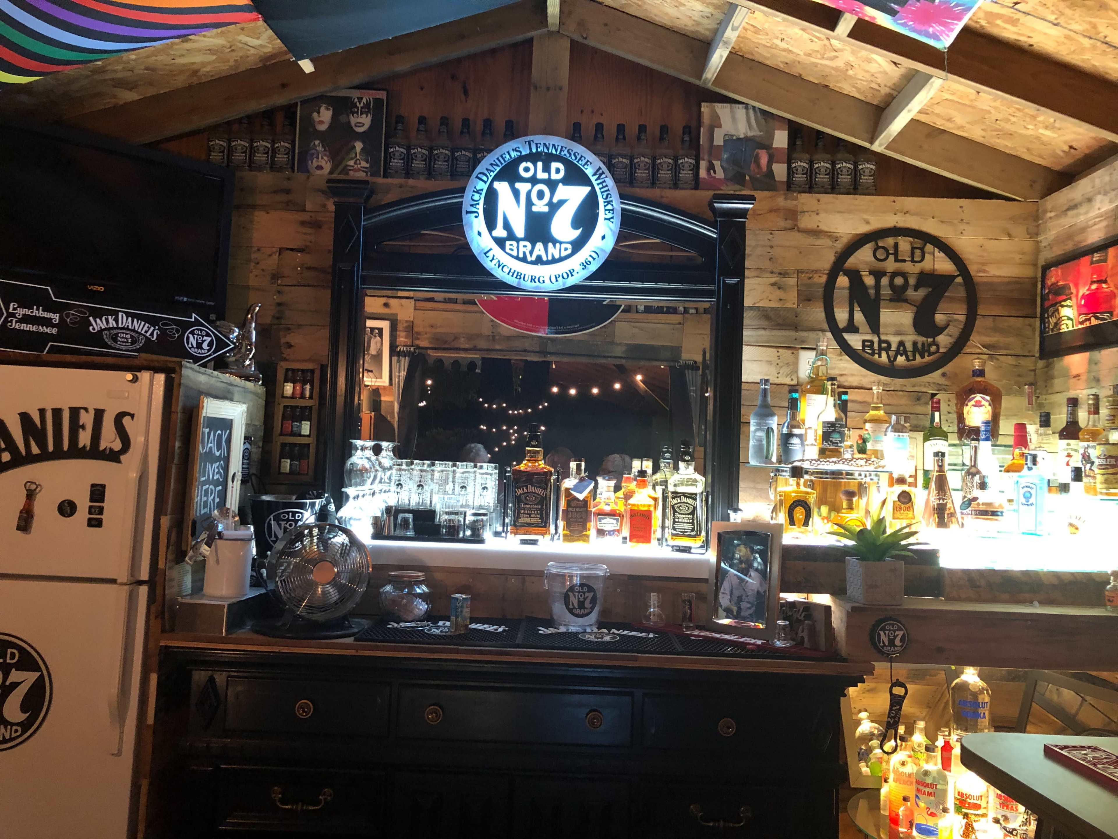 A rustic bar area with a selection of whiskey bottles displayed behind a counter, illuminated by a glowing round sign that reads "Old No. 7 Brand."
