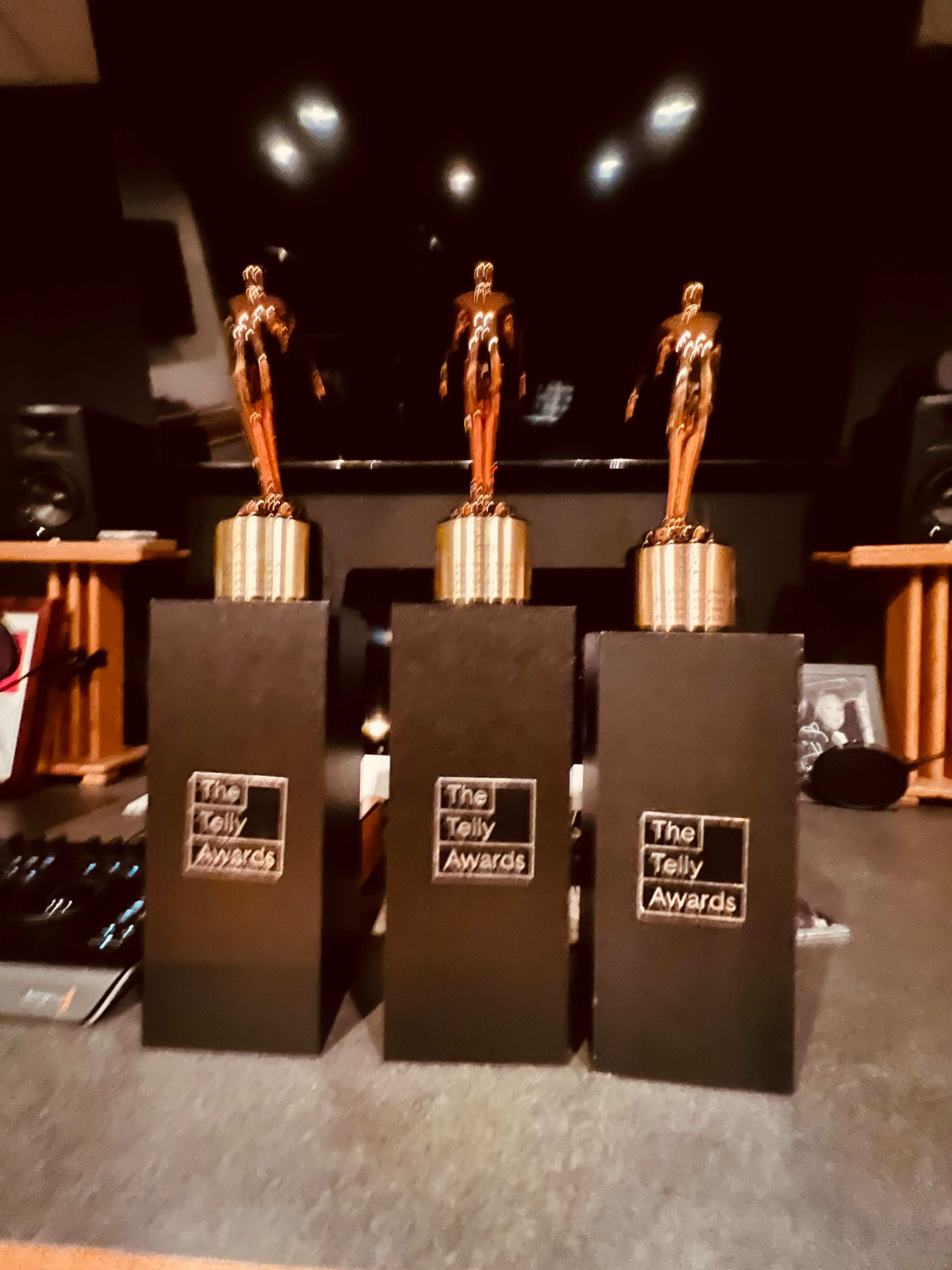 Three Telly Award trophies are displayed on a table in front of a dark background with speakers.