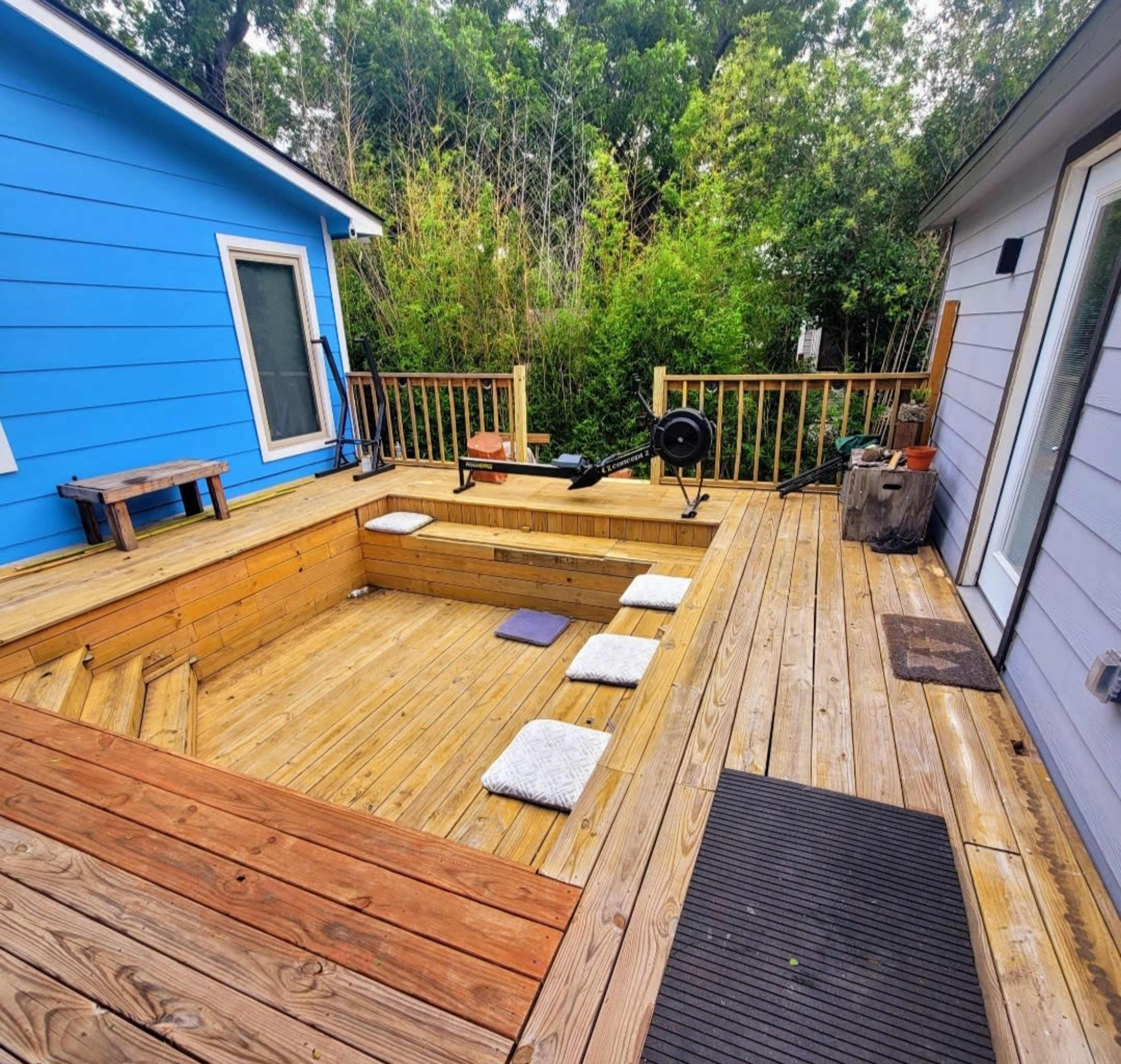 The image shows a wooden deck with a sunken area, surrounded by a railing and several workout equipment pieces, set against a backdrop of greenery.