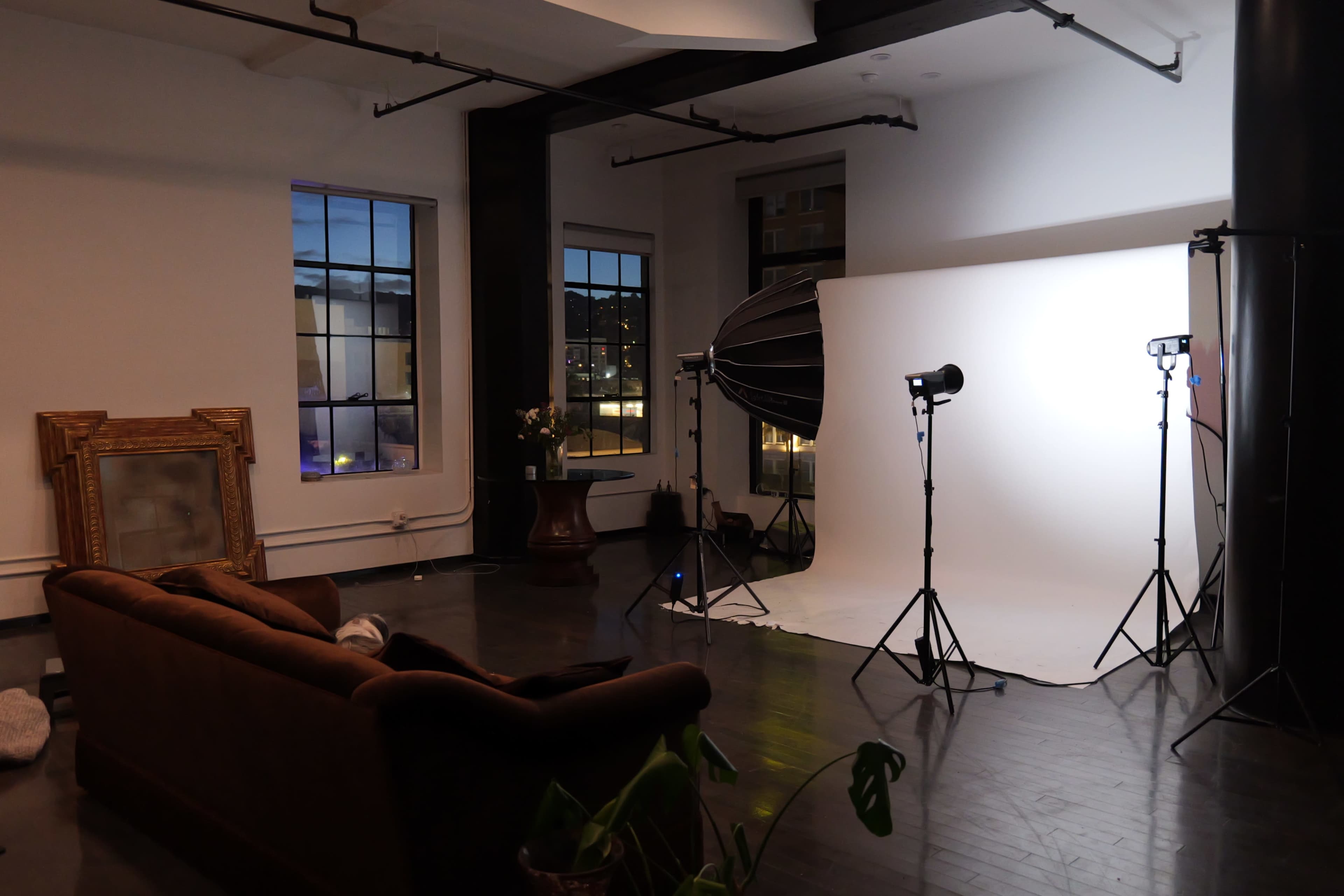 The image shows a studio space with a brown sofa, large windows, a lighting setup, and a backdrop for photography.