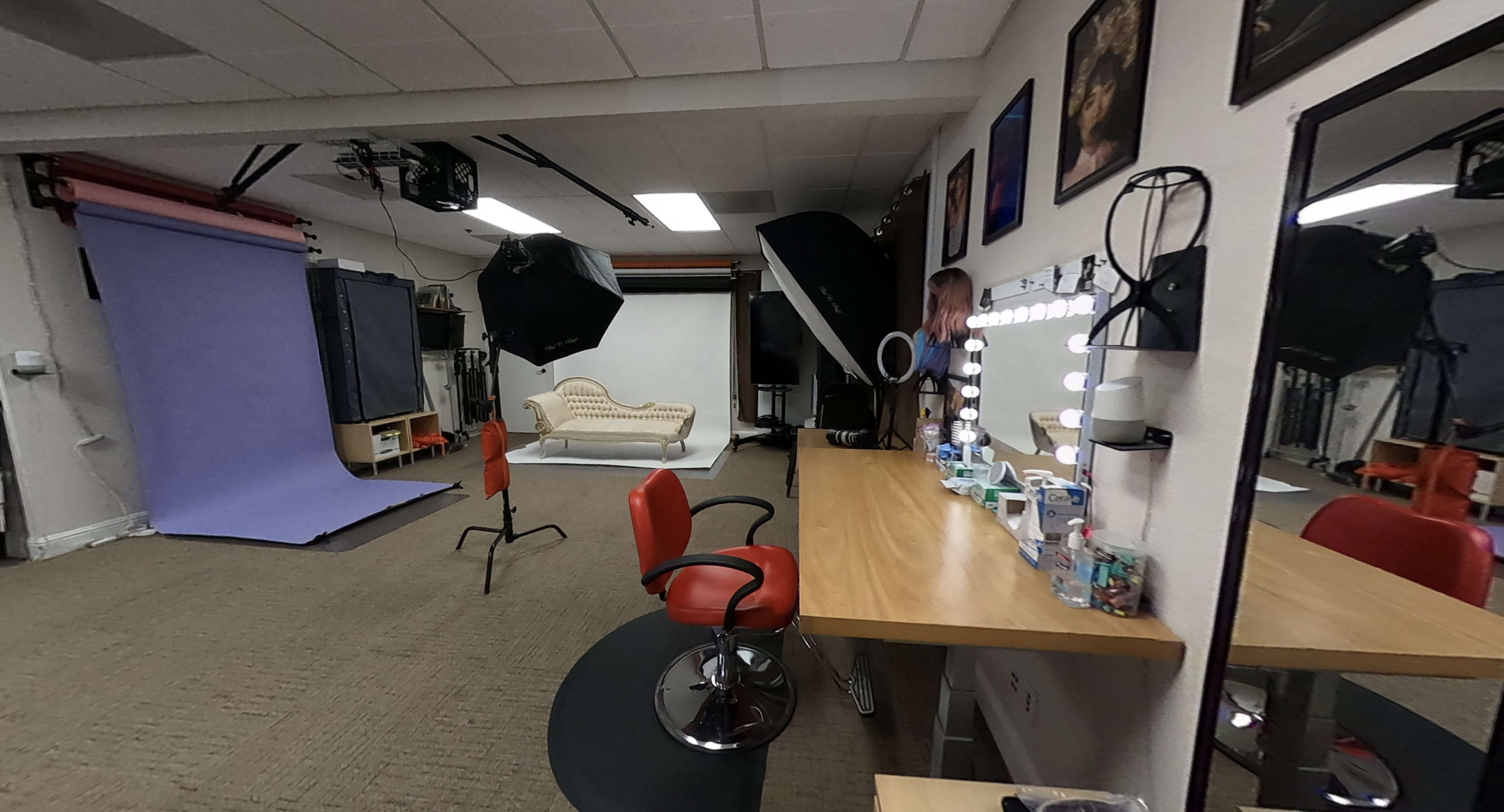 A photography studio with a lighted vanity mirror, a red chair, a beige couch, and equipment set up against a backdrop.