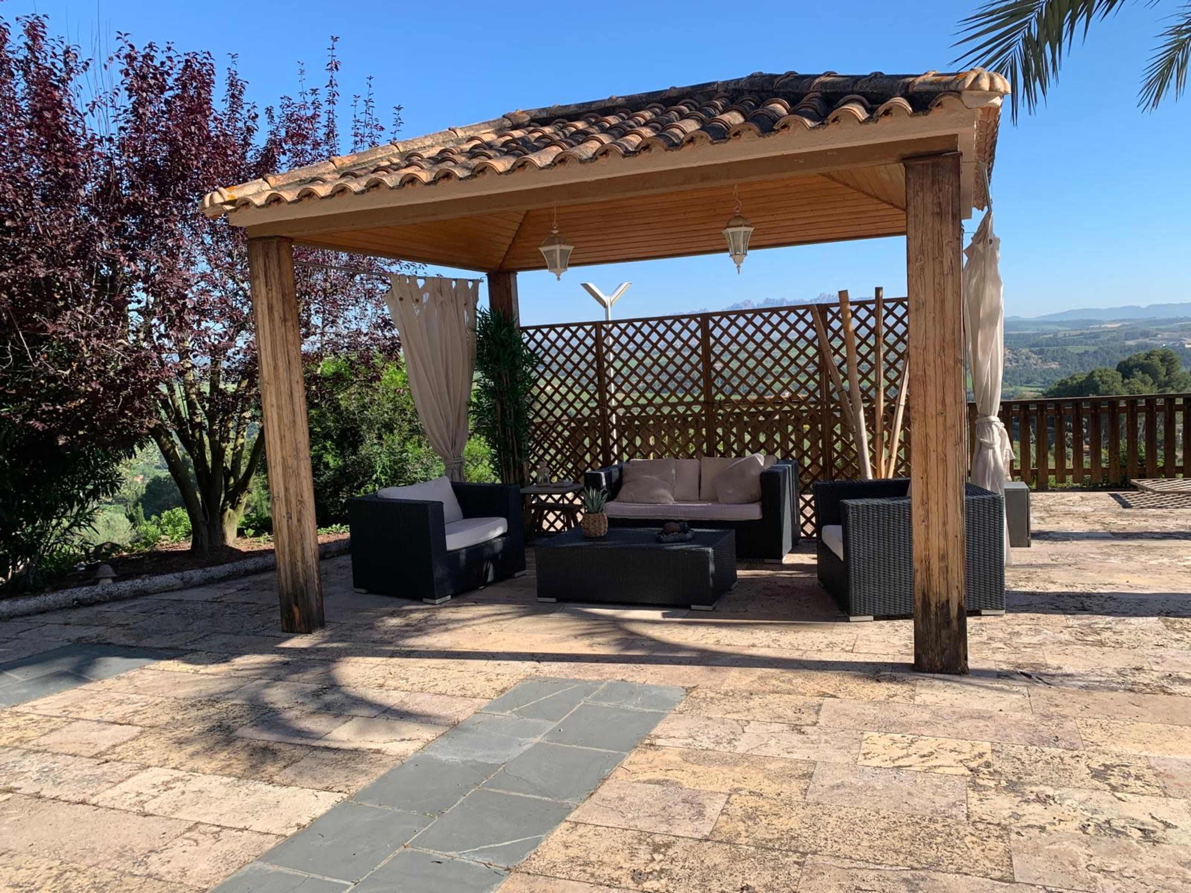 The image shows a shaded outdoor seating area with a tiled floor, decorative wooden beams, and comfortable furniture surrounded by greenery.