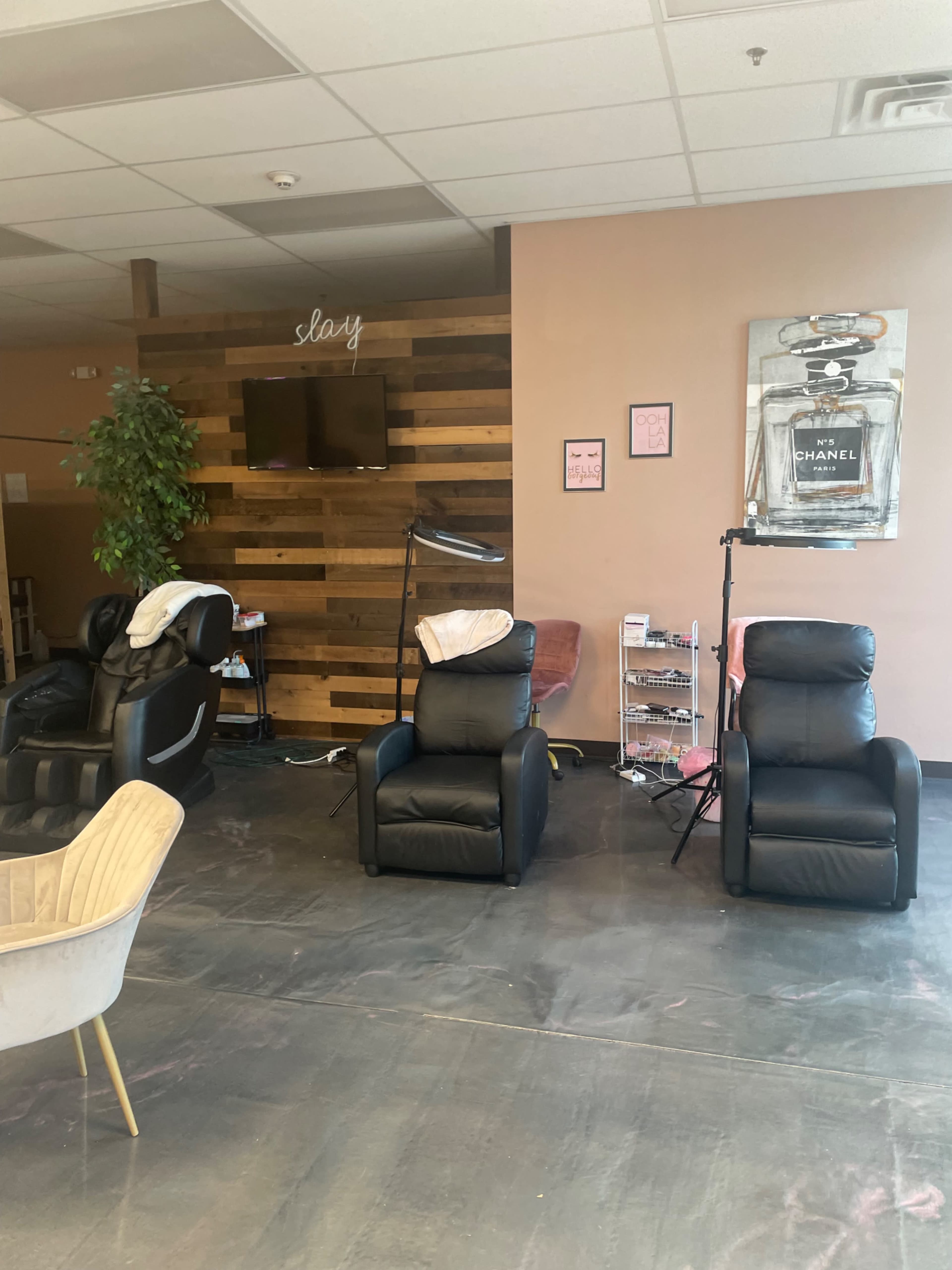 A beauty salon interior with three black reclining chairs, a wood-paneled wall, a television, and various salon equipment.