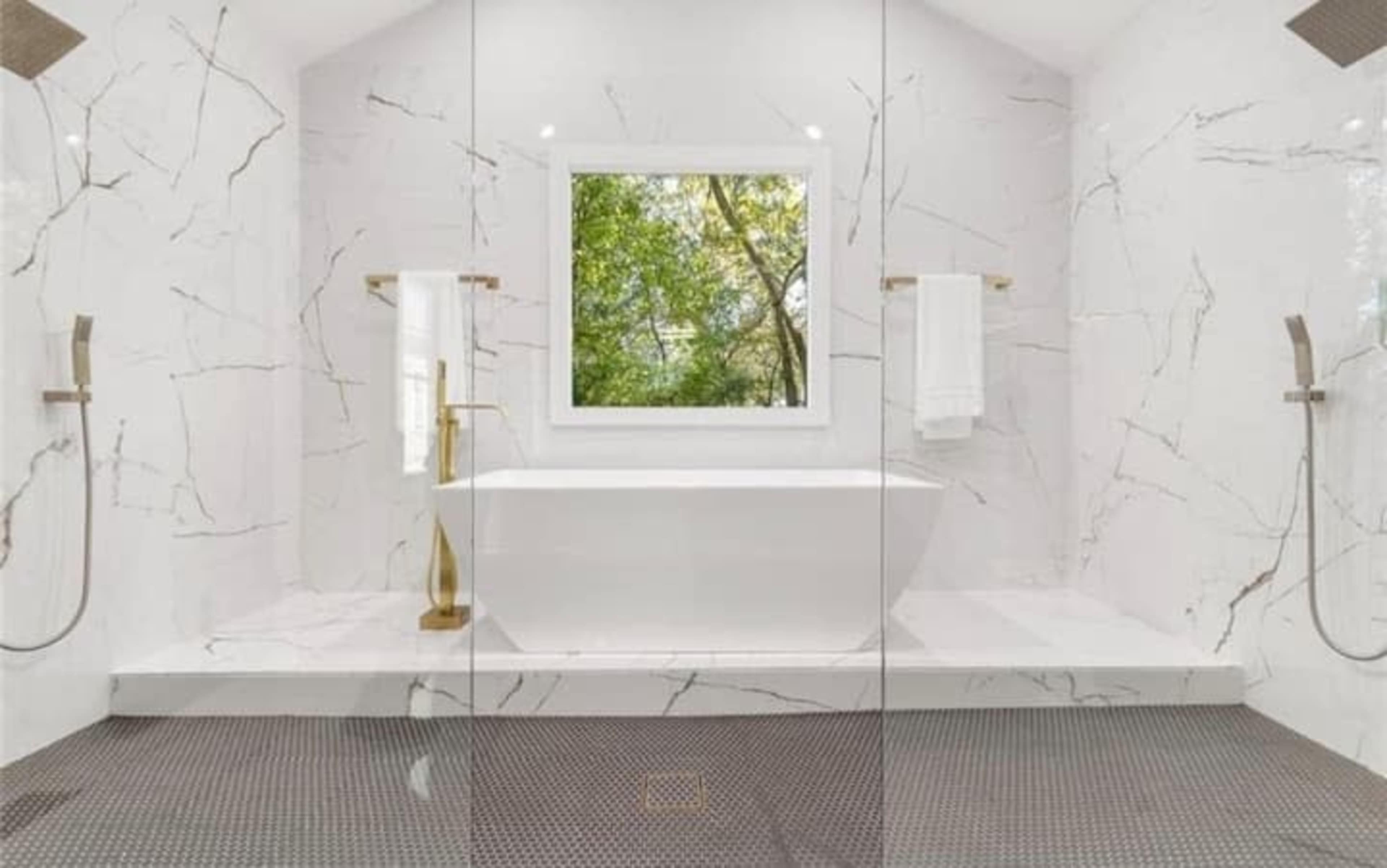 A modern bathroom featuring a freestanding bathtub, large windows with a view of greenery, and marble walls with gold fixtures.