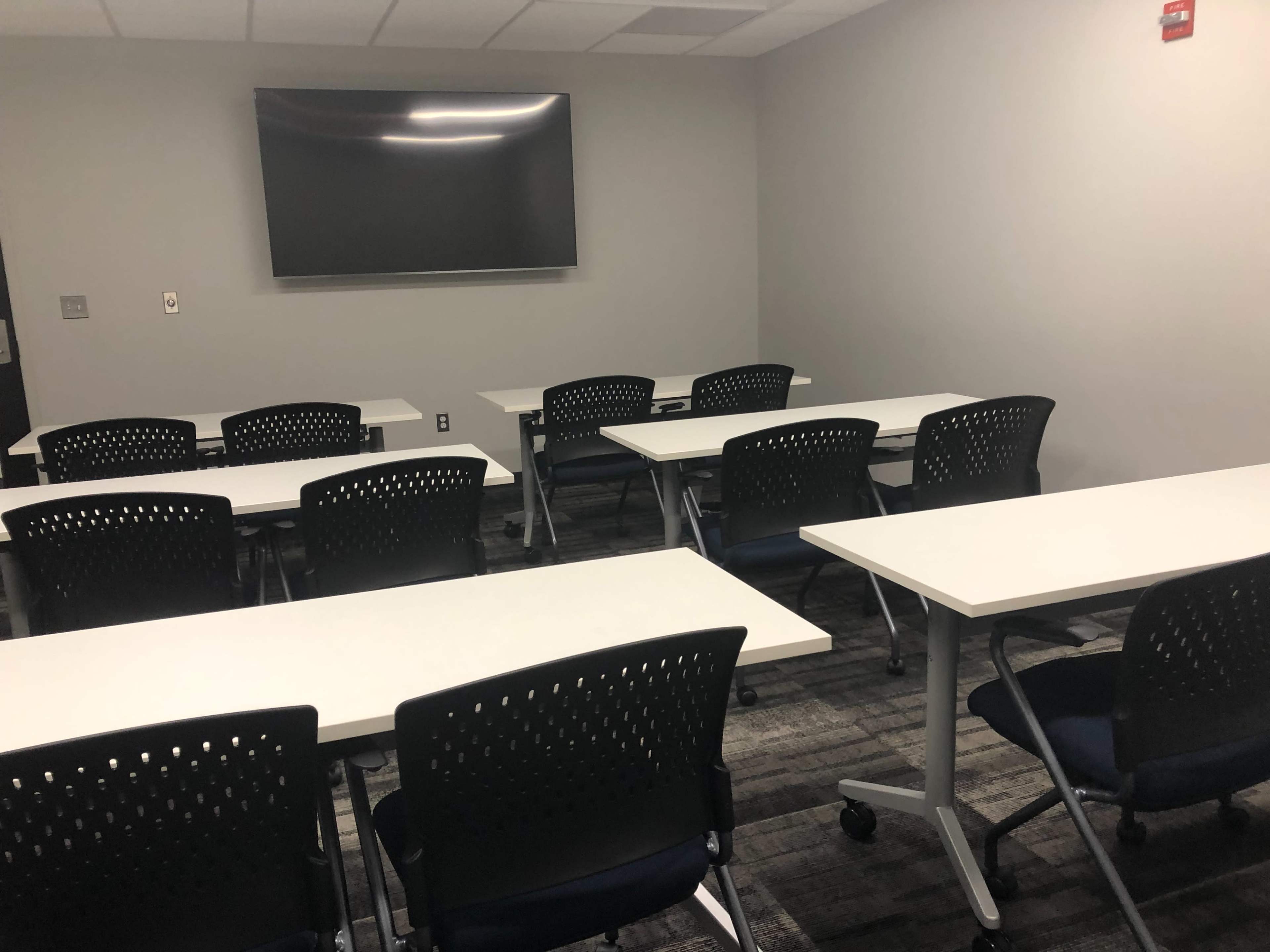 A modern classroom with several white tables and black chairs arranged in rows, facing a wall-mounted screen.