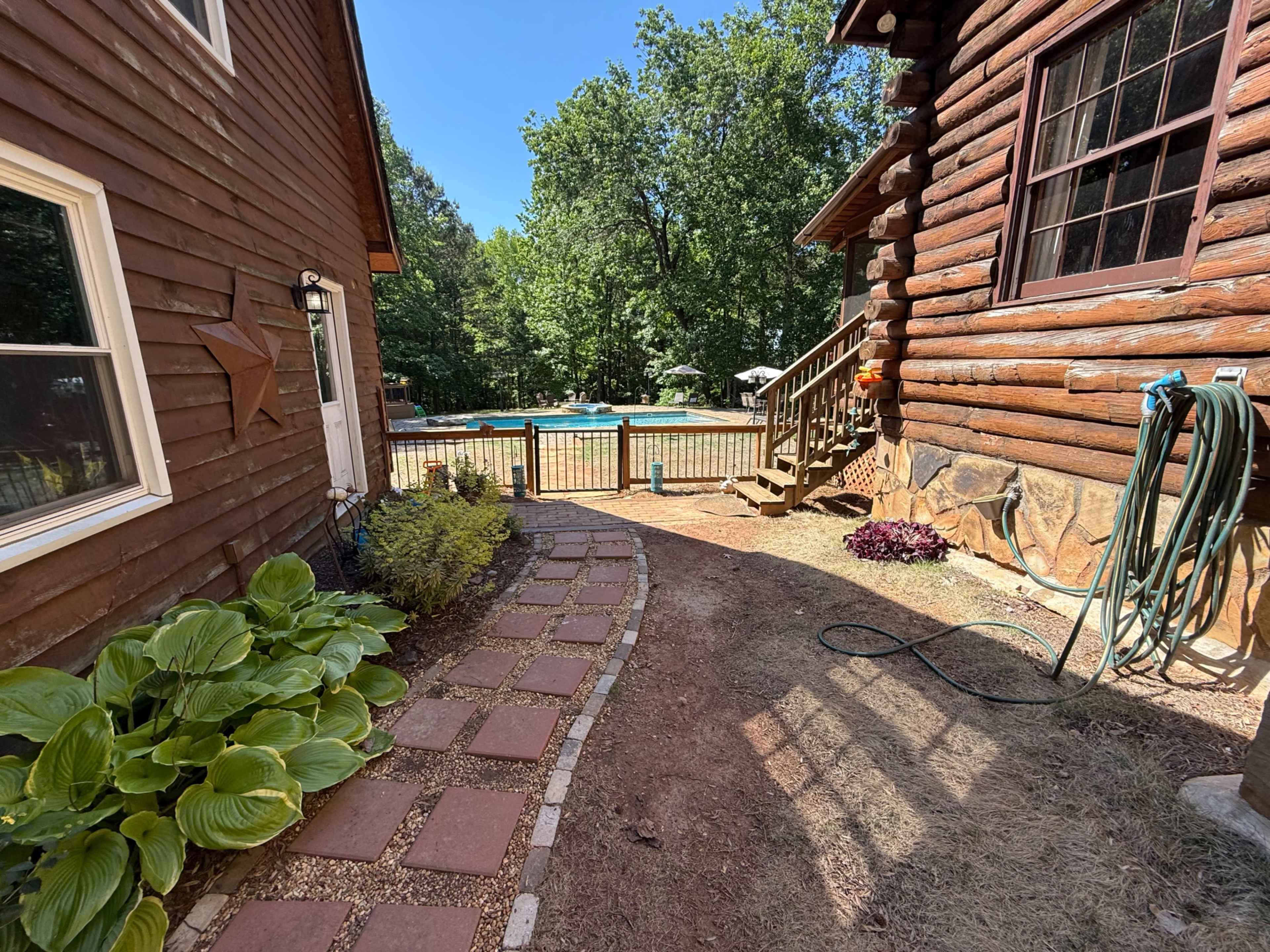 The image shows a narrow pathway lined with brick pavers leading to a fenced area with a swimming pool, bordered by wooden structures and greenery.