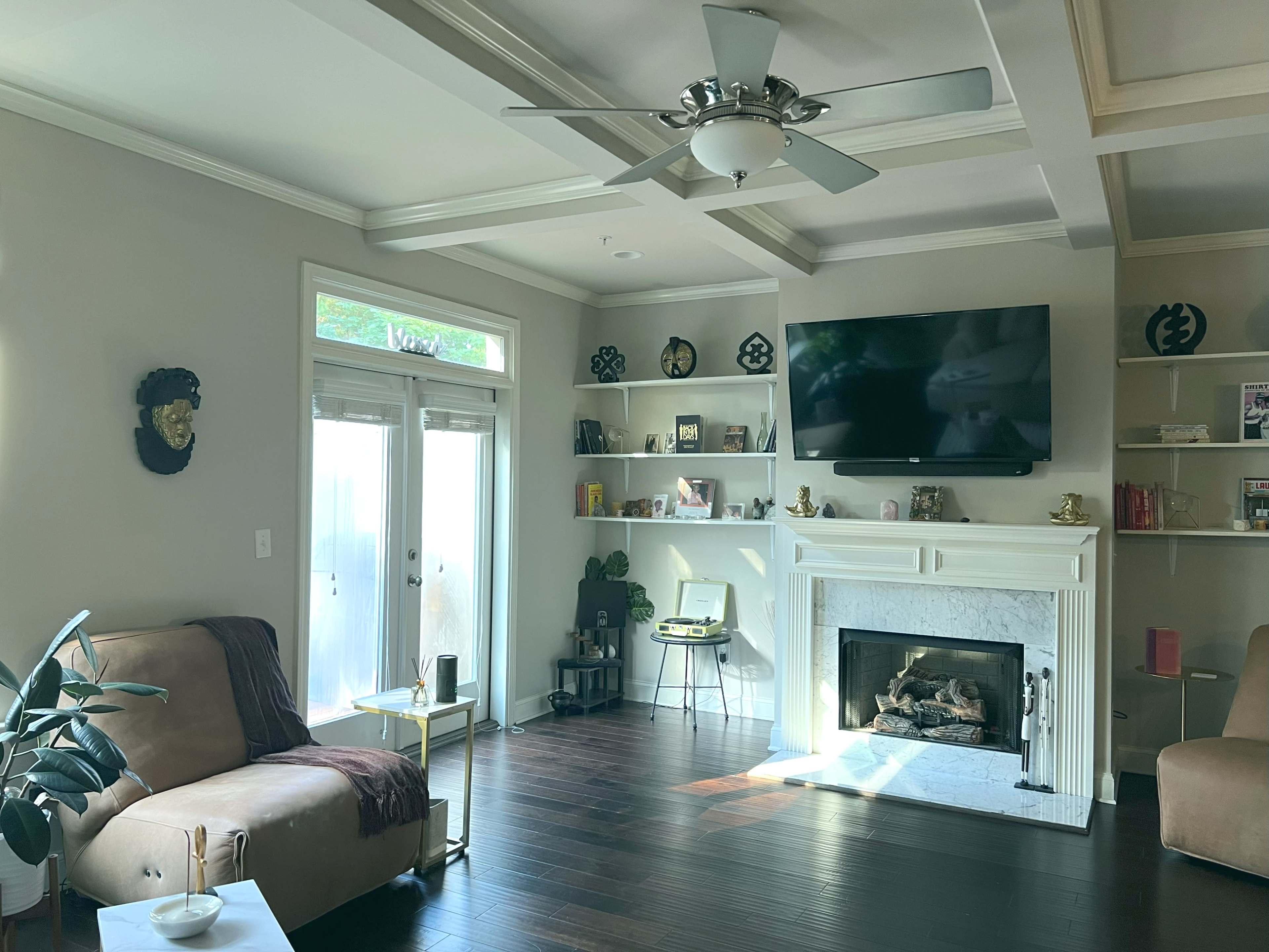 The living room features a sectional sofa, a coffee table, a fireplace, and wall shelves with decorative items, illuminated by natural light from the windows.
