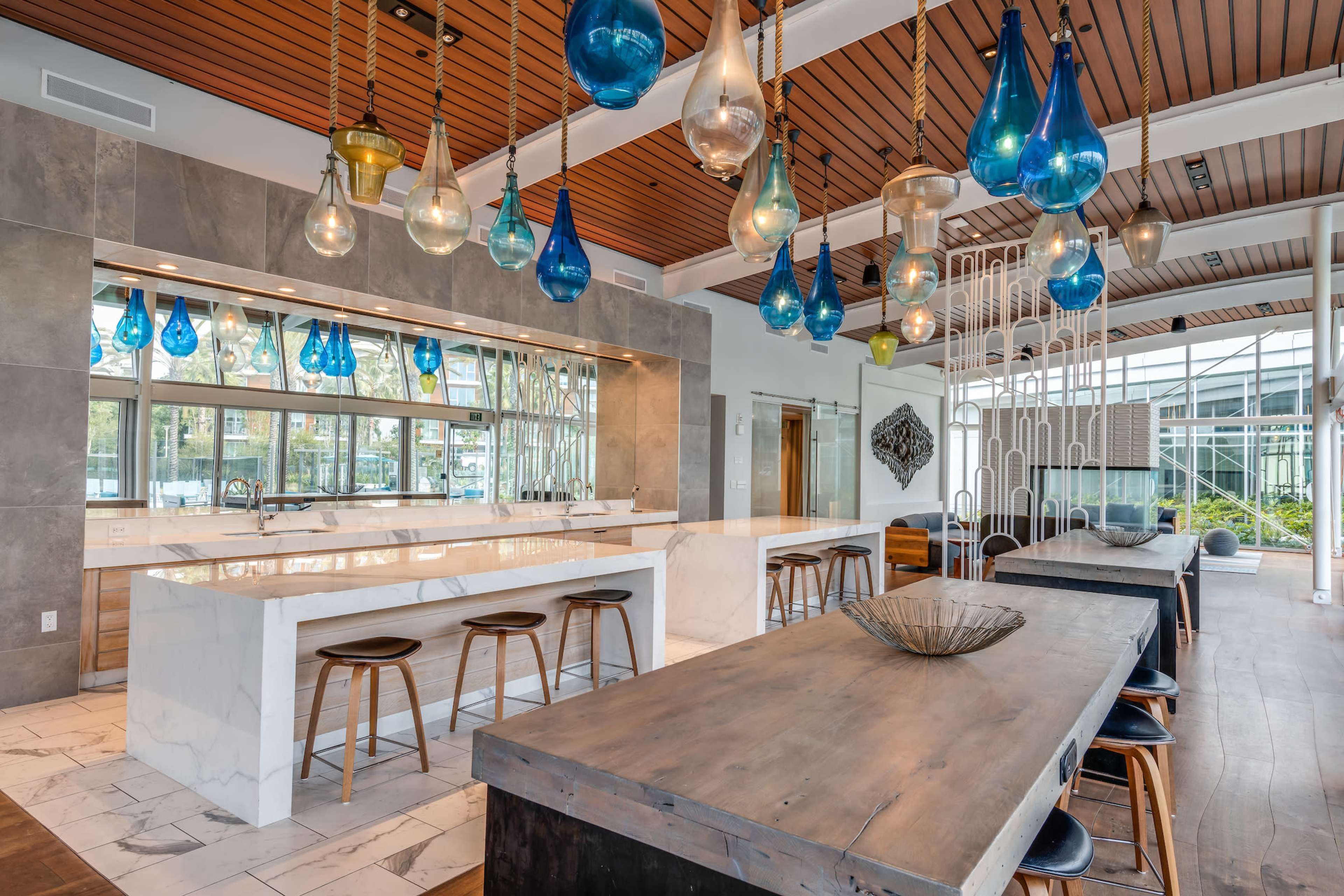 A modern kitchen features white marble countertops, wooden stools, and colorful pendant lights hanging from a wooden ceiling.