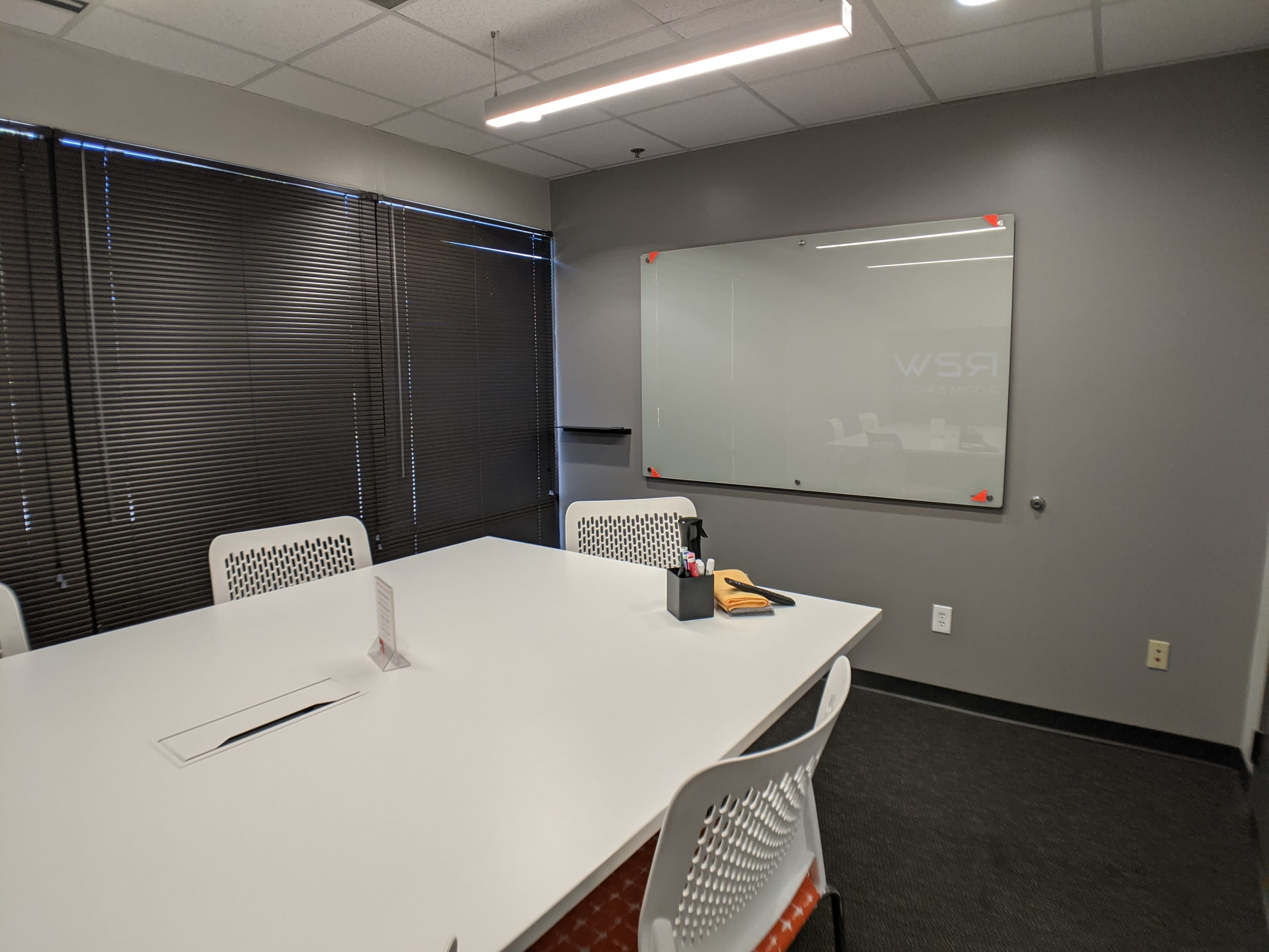 A conference room with a large white table, several white chairs, and a glass board on the wall.