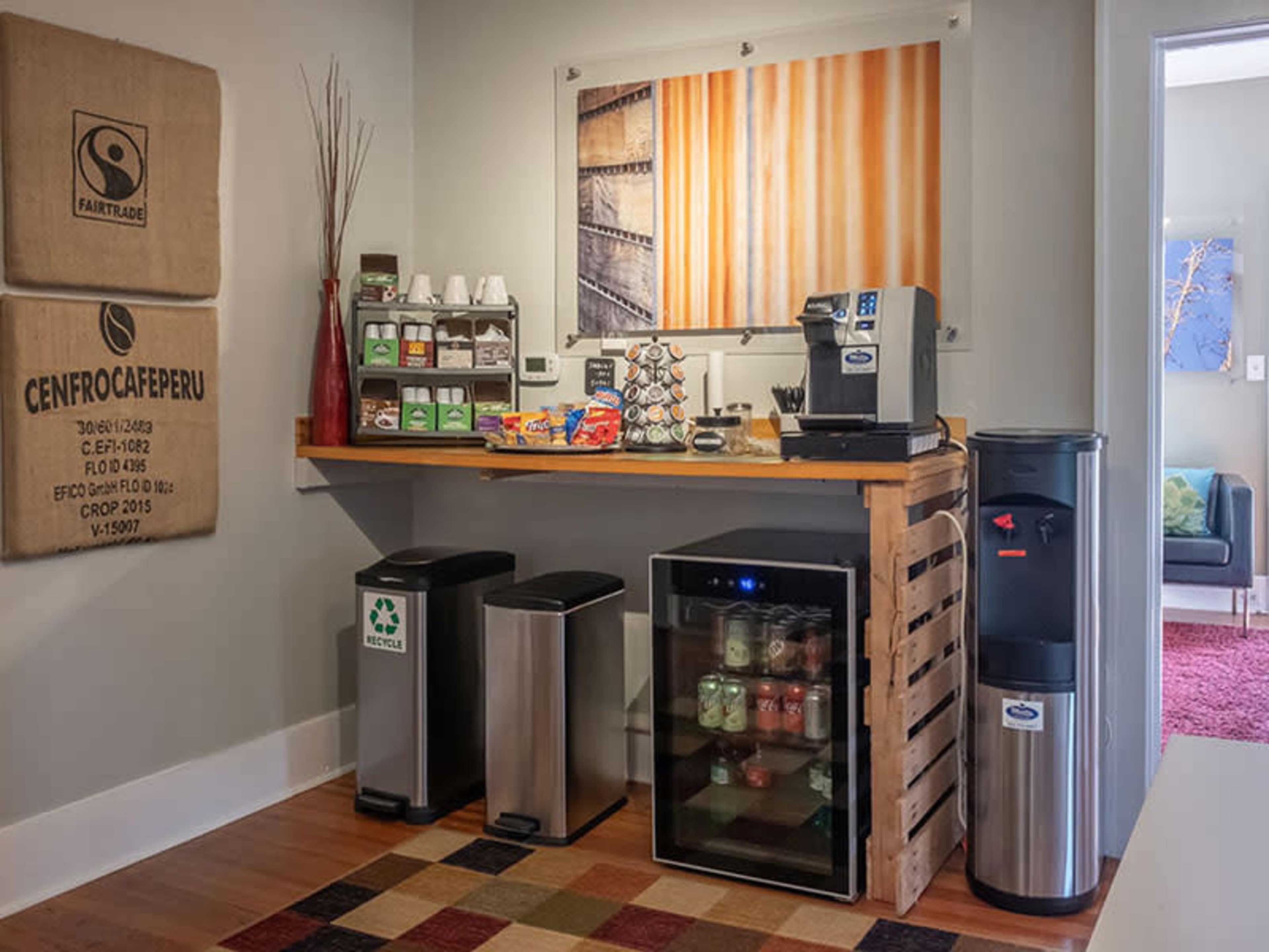 A small kitchen area with a coffee machine, snack items, a recycling bin, and a stocked refrigerator against a backdrop of wooden and decorative wall art.