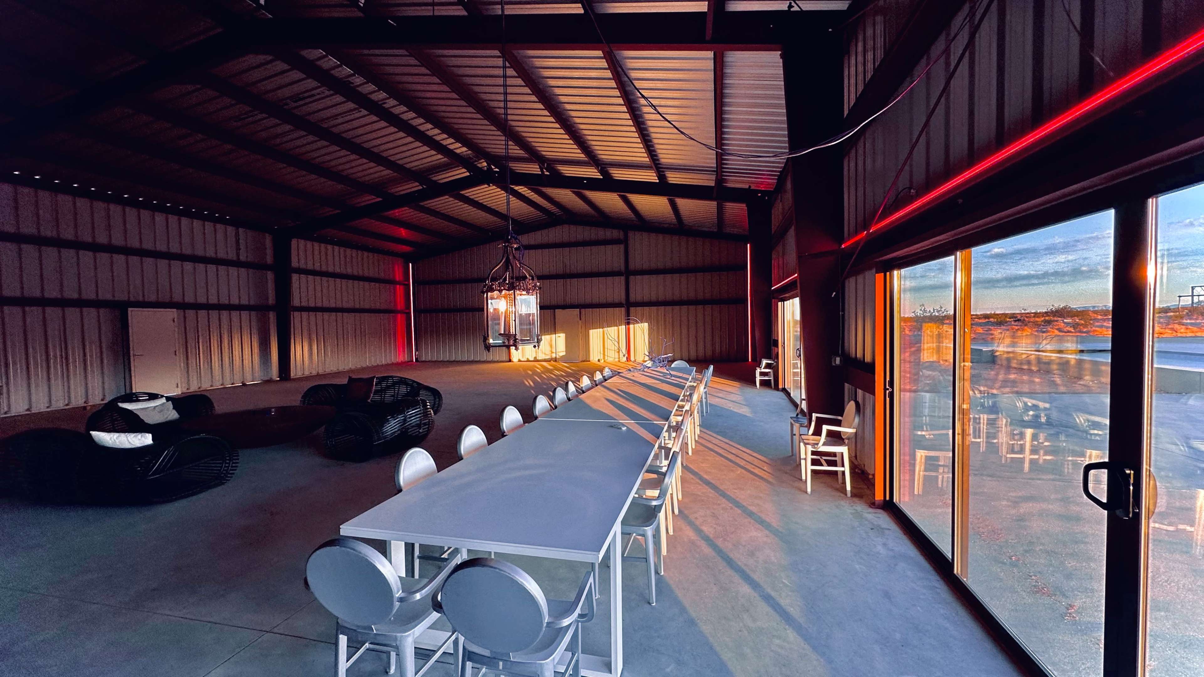 The image shows a spacious, industrial-style interior with a long dining table, modern seating, and large glass doors leading to an outdoor view.