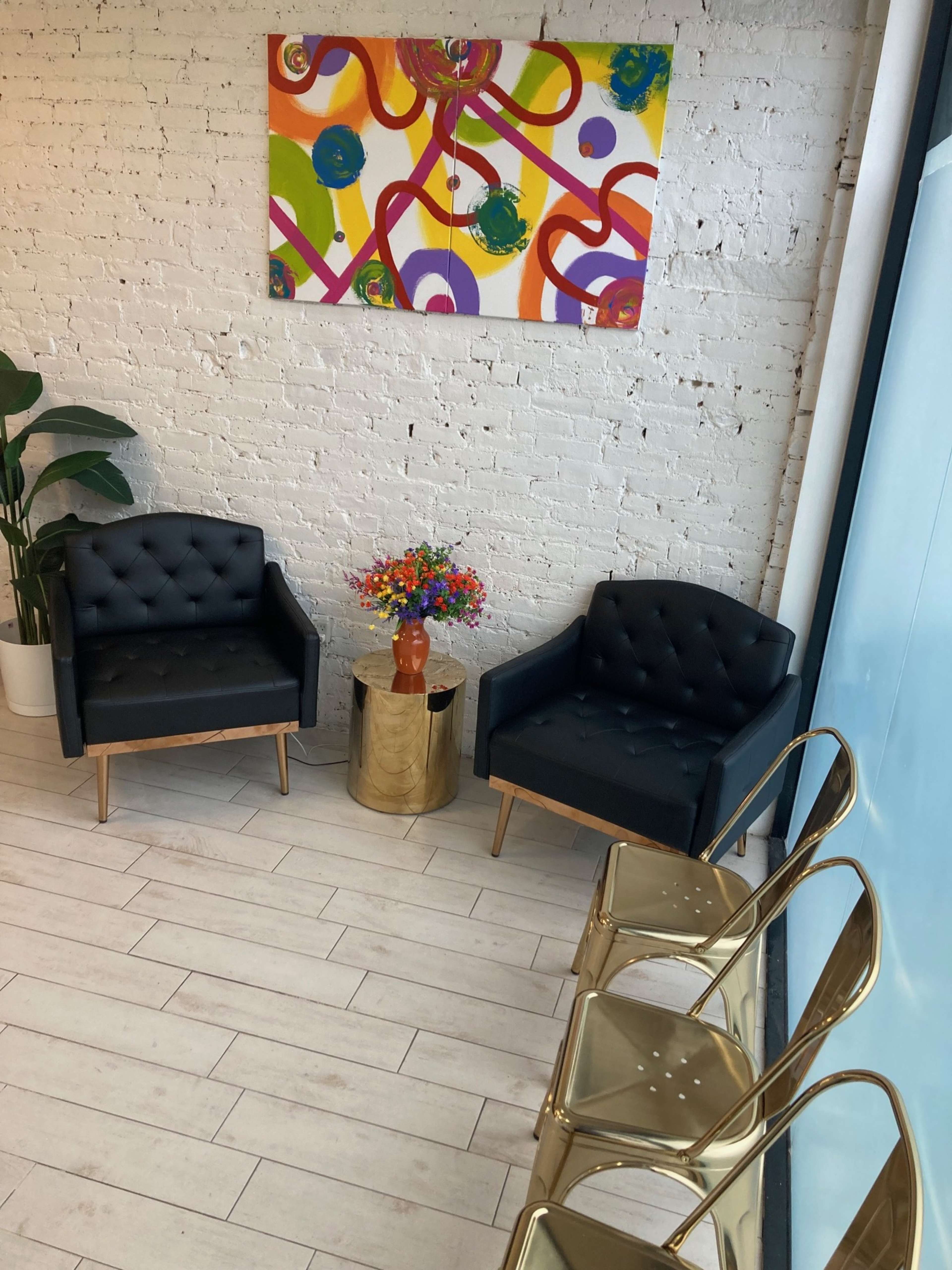 The image shows a waiting area with two black tufted chairs, a colorful abstract painting on the wall, a round gold table with a floral arrangement, and metal chairs nearby.