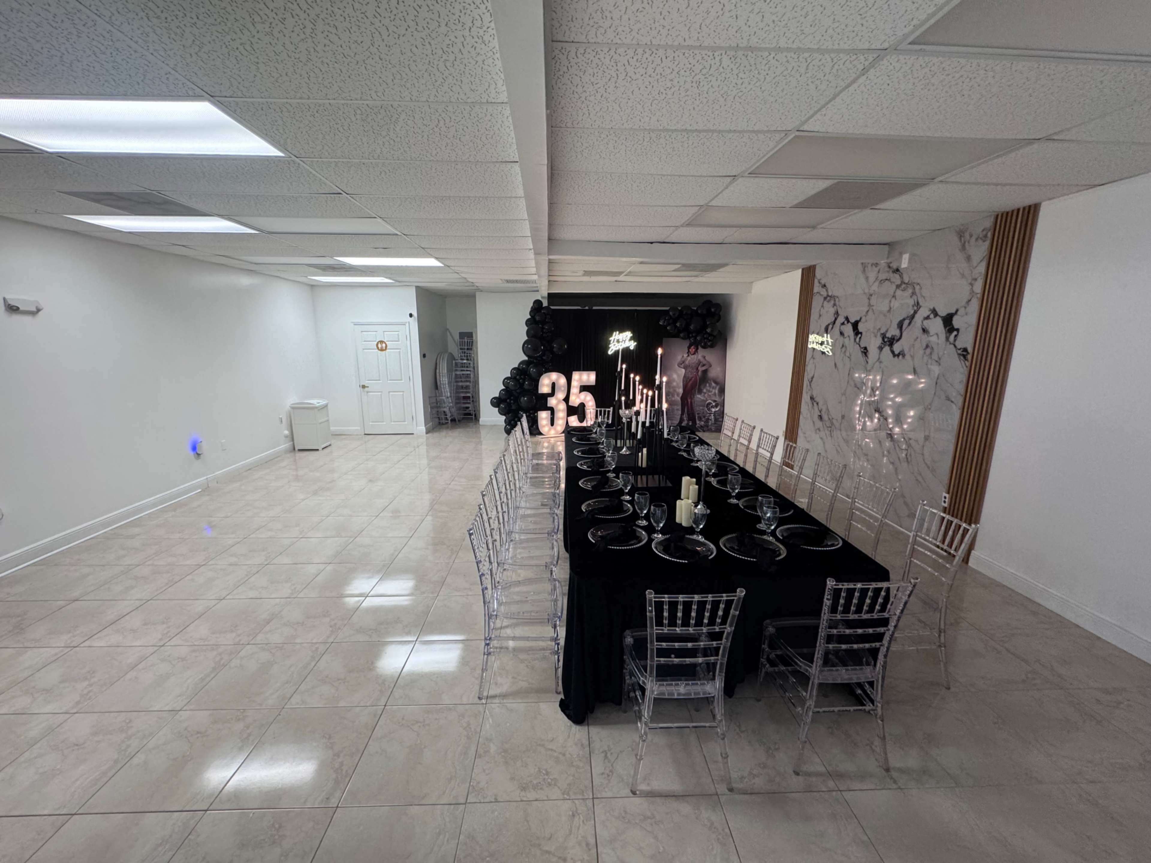 A long black table with place settings is arranged in a spacious, well-lit room decorated with black balloons and illuminated numbers prominently displaying "35."