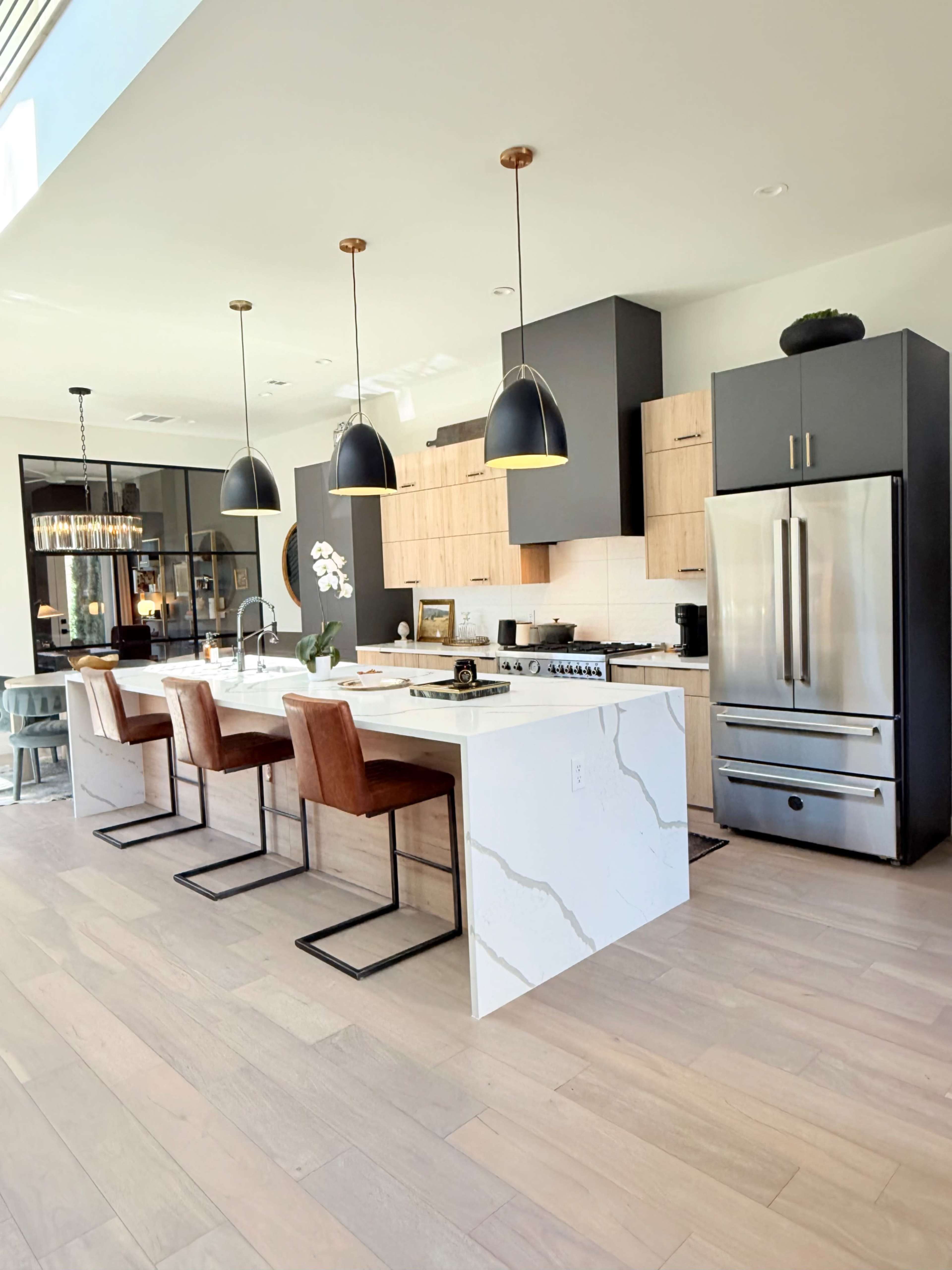 The image shows a modern kitchen with a large island featuring three bar stools, pendant lights overhead, and stainless steel appliances in a light wood and grey cabinet setup.