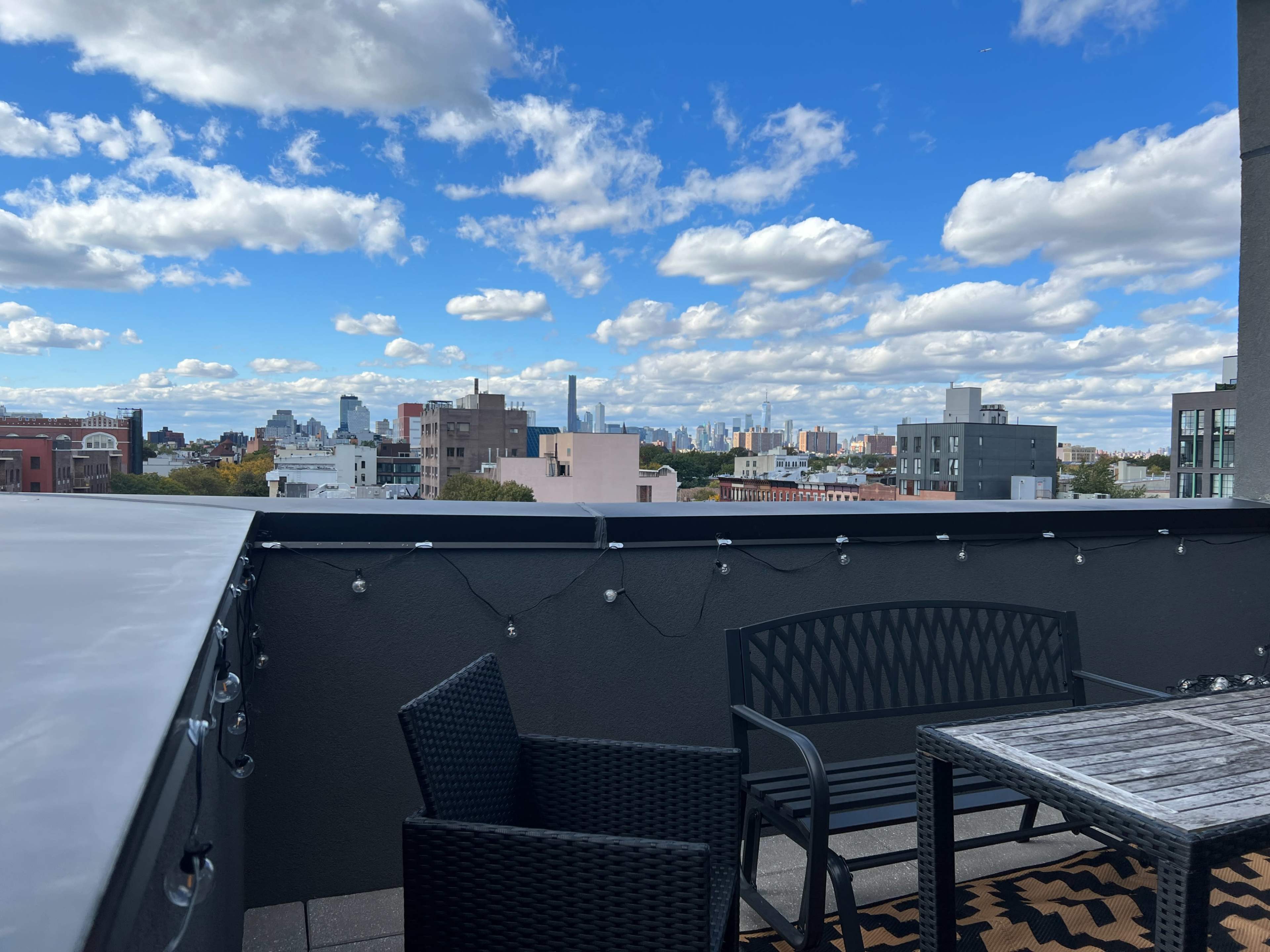A rooftop terrace features a table and chairs with a city skyline and blue sky adorned with clouds in the background.