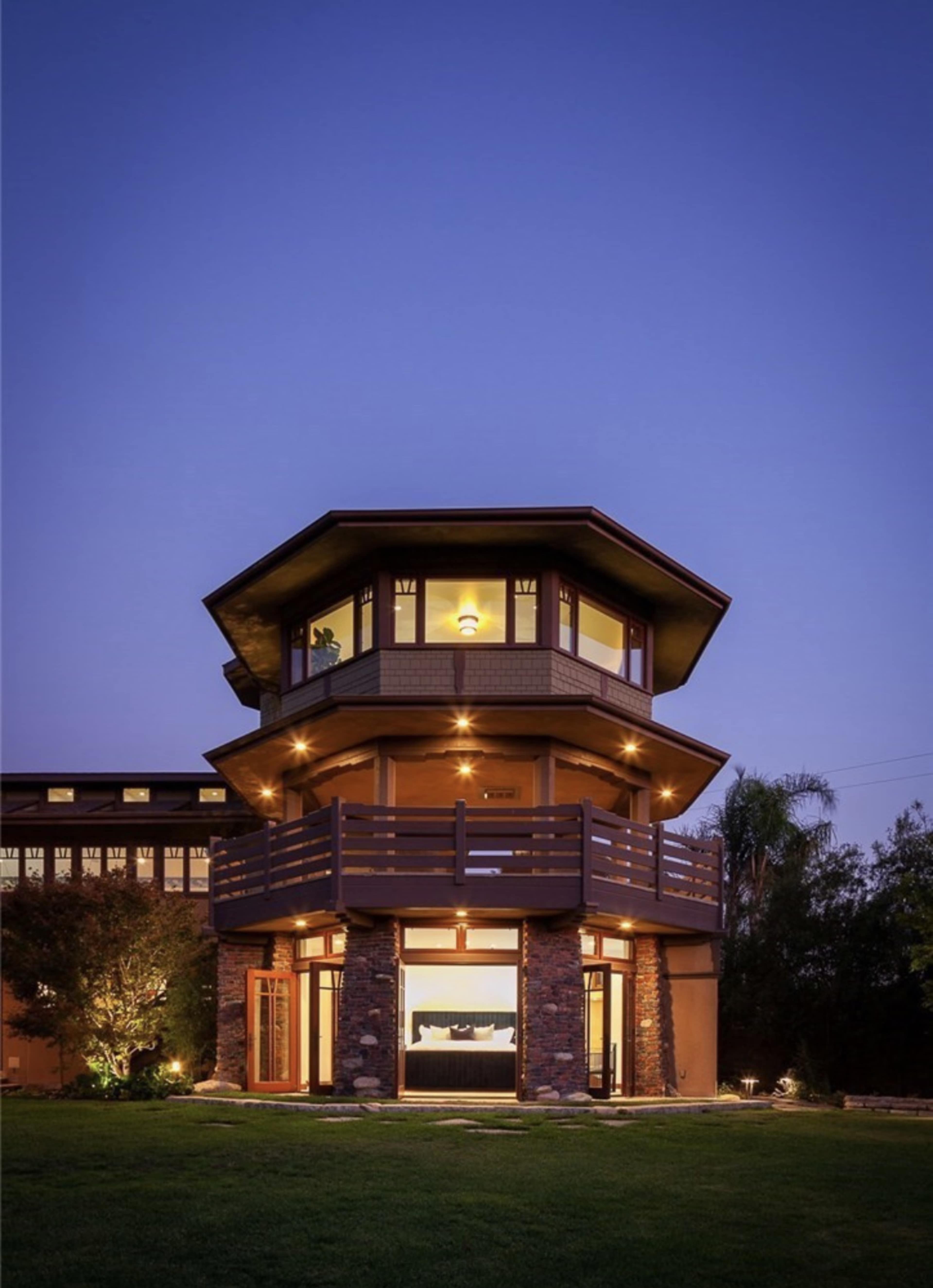 A multi-story, modern house with a circular design features large windows, wooden balconies, and stone accents, set against a twilight sky.