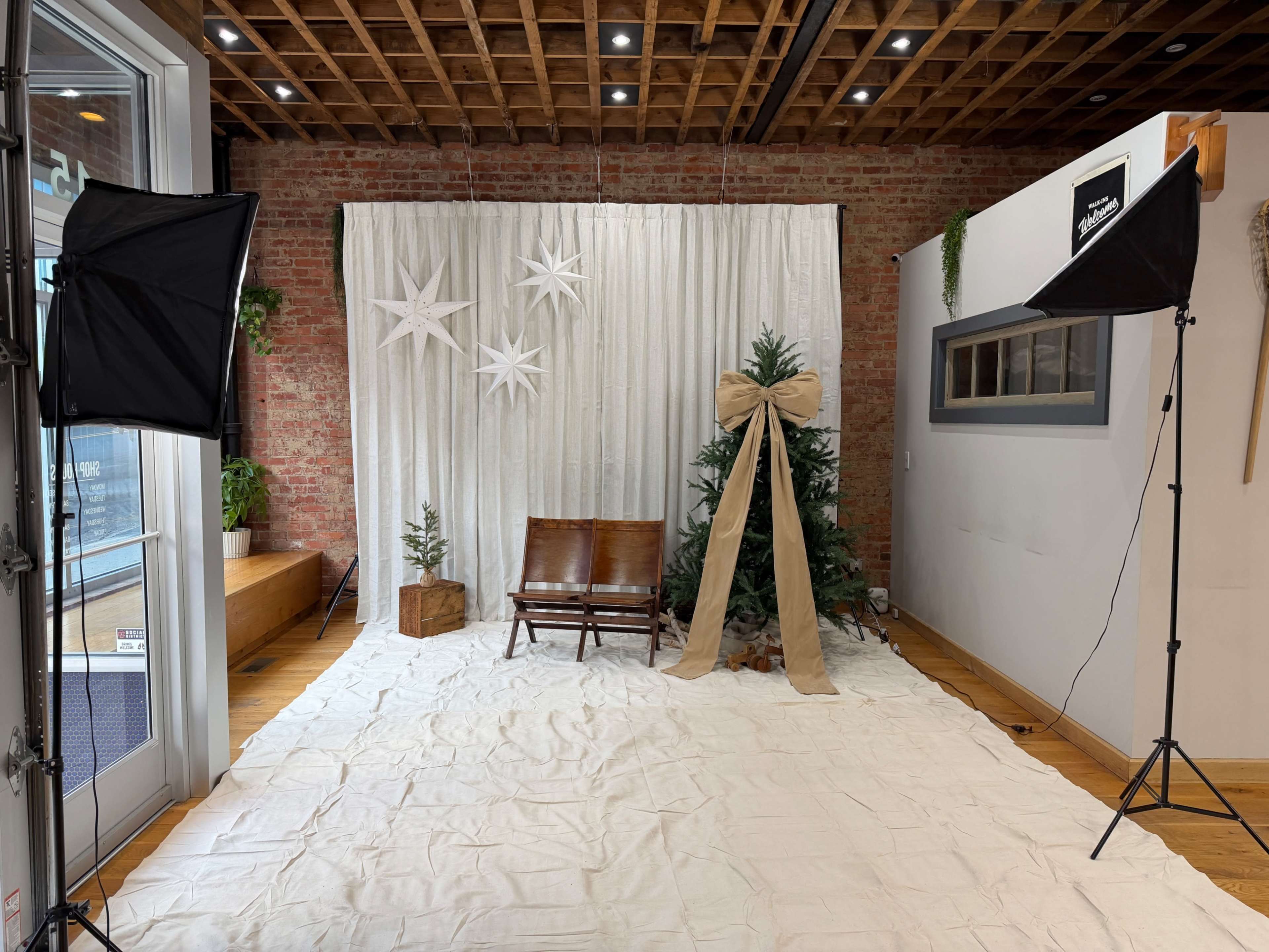 The image shows a photo backdrop setup featuring a white curtain with decorative stars, a Christmas tree adorned with a large bow, and two softbox lights on either side, arranged on a white fabric-covered floor.