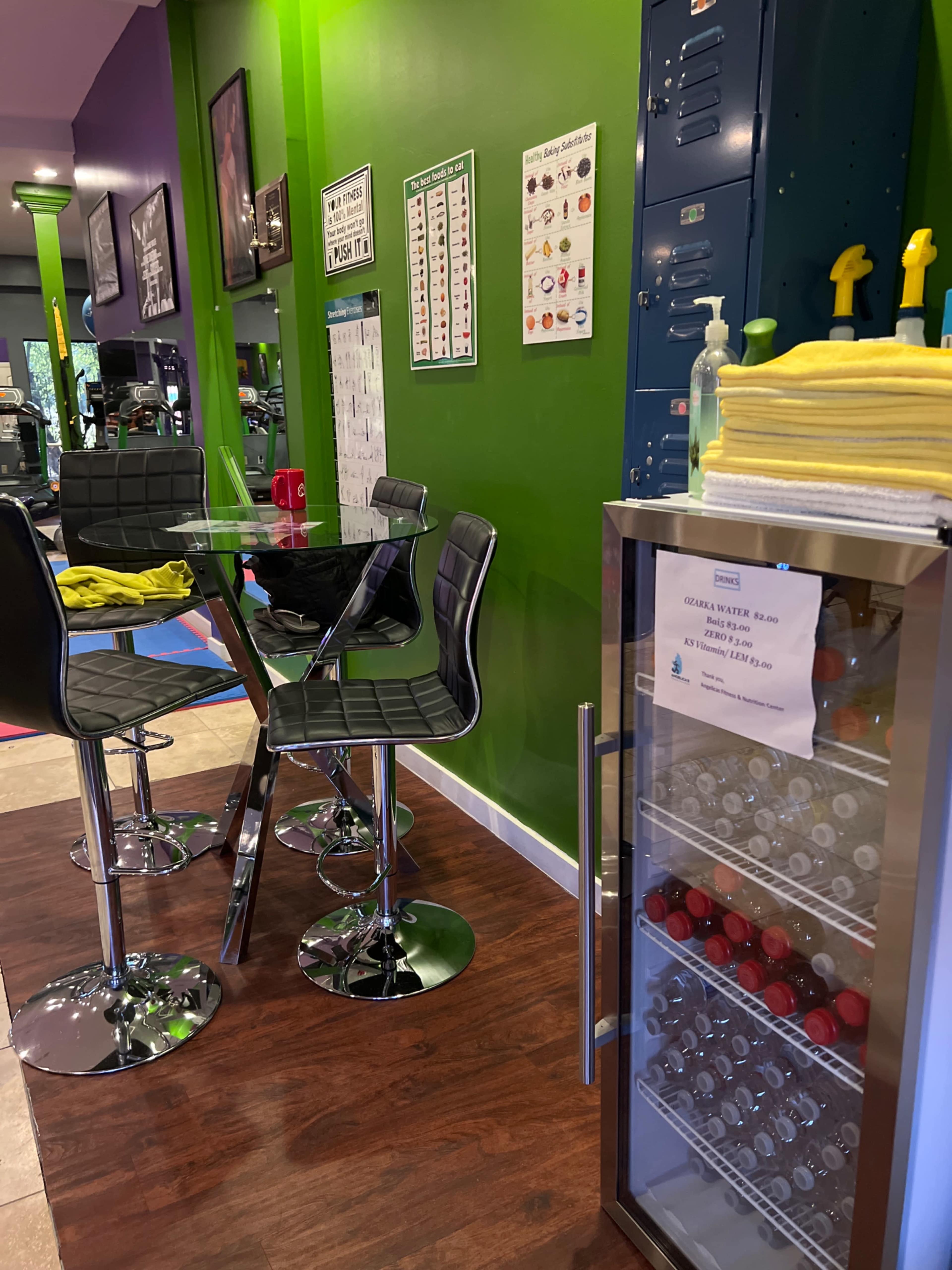 The image shows a small lounge area in a gym featuring a glass table with black chairs, a green wall, and a refrigerated display case filled with drinks.