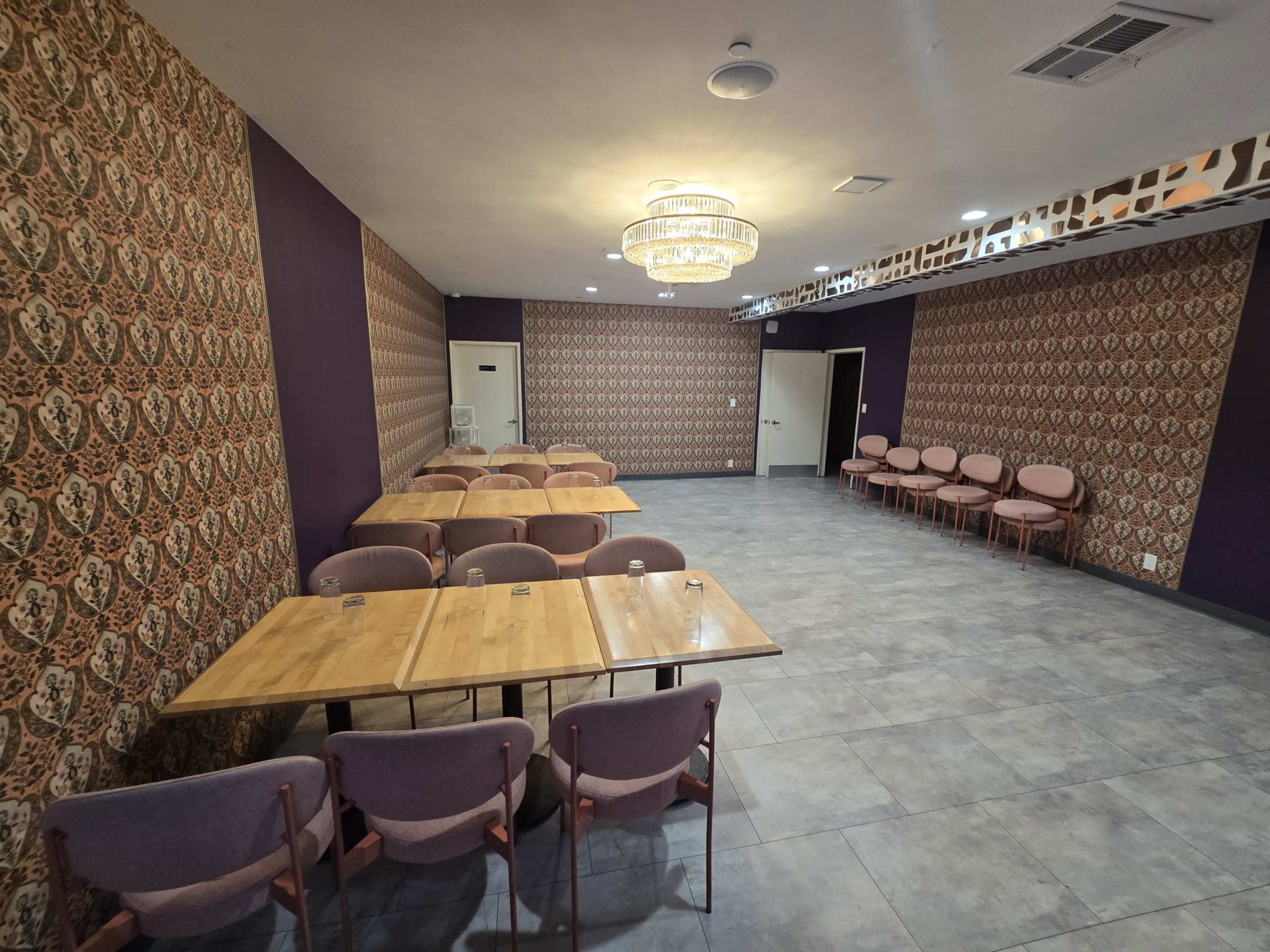 The image shows a spacious room with patterned wallpaper, a chandelier, and several tables arranged for seating.