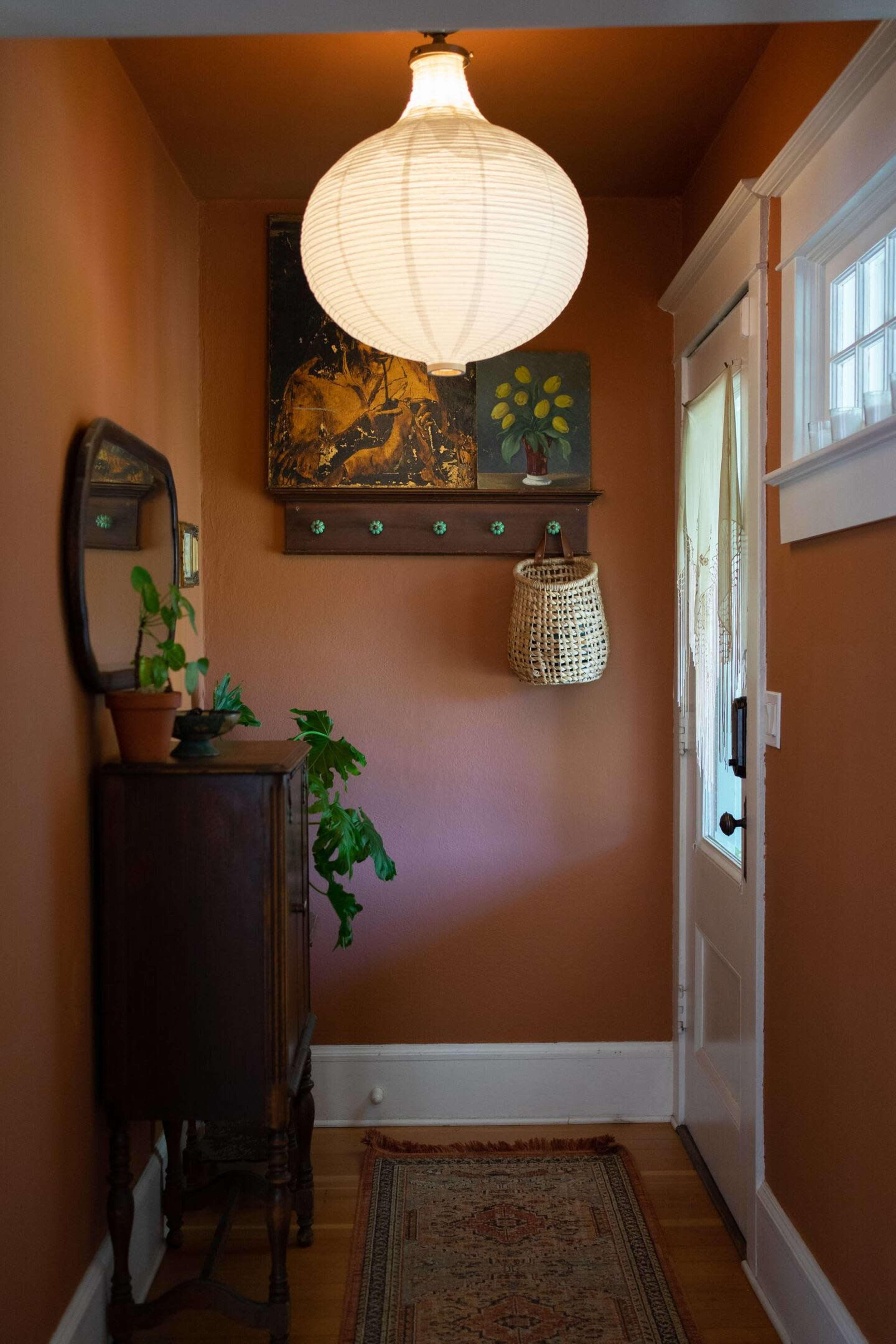 A hallway with an orange wall, a round pendant lamp, a small wooden cabinet, a mirror, a woven basket, and a door leading outside.