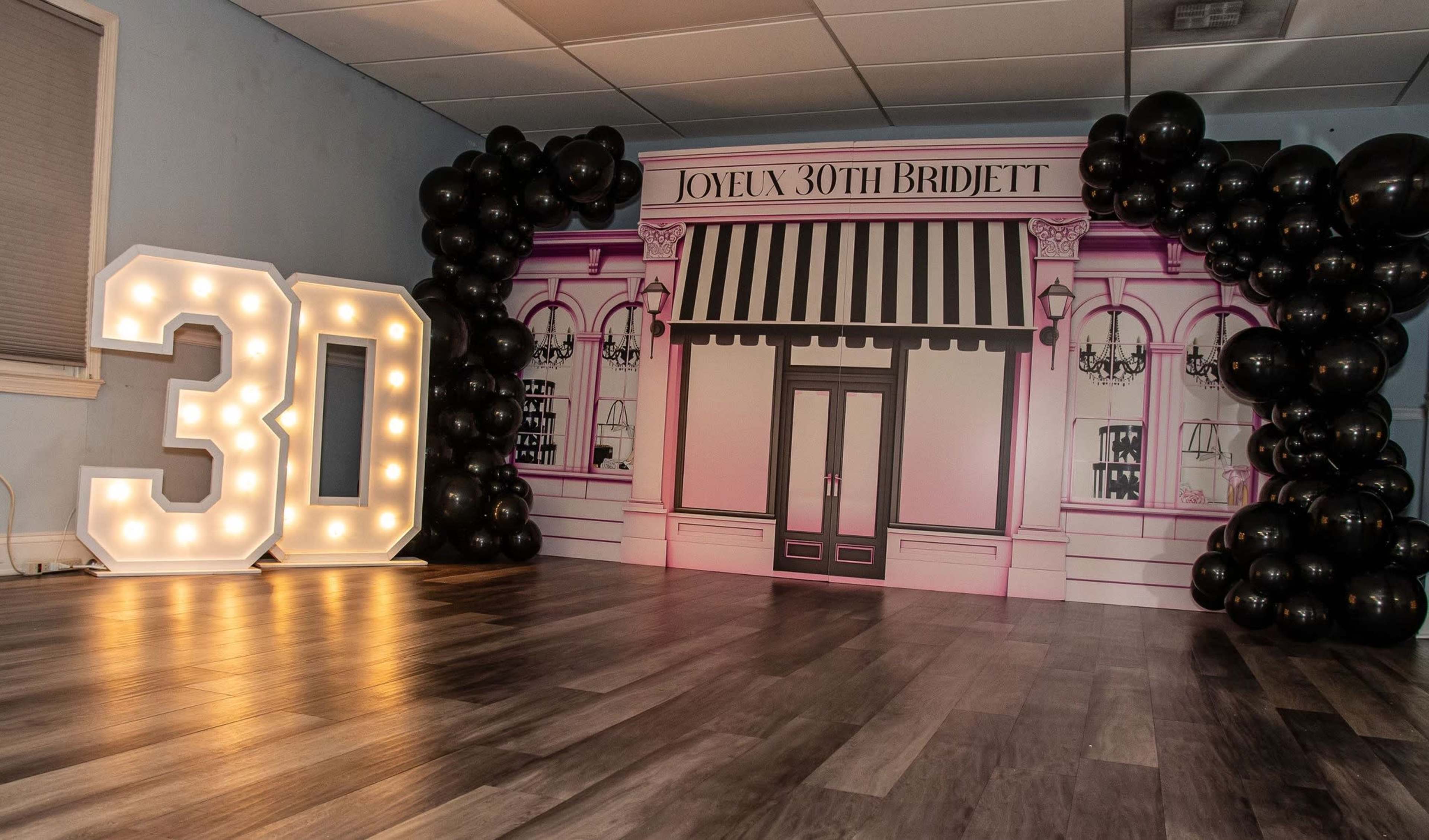 A decorated indoor space featuring a large backdrop with a storefront design and oversized illuminated numbers "30" next to a cluster of black balloons.
