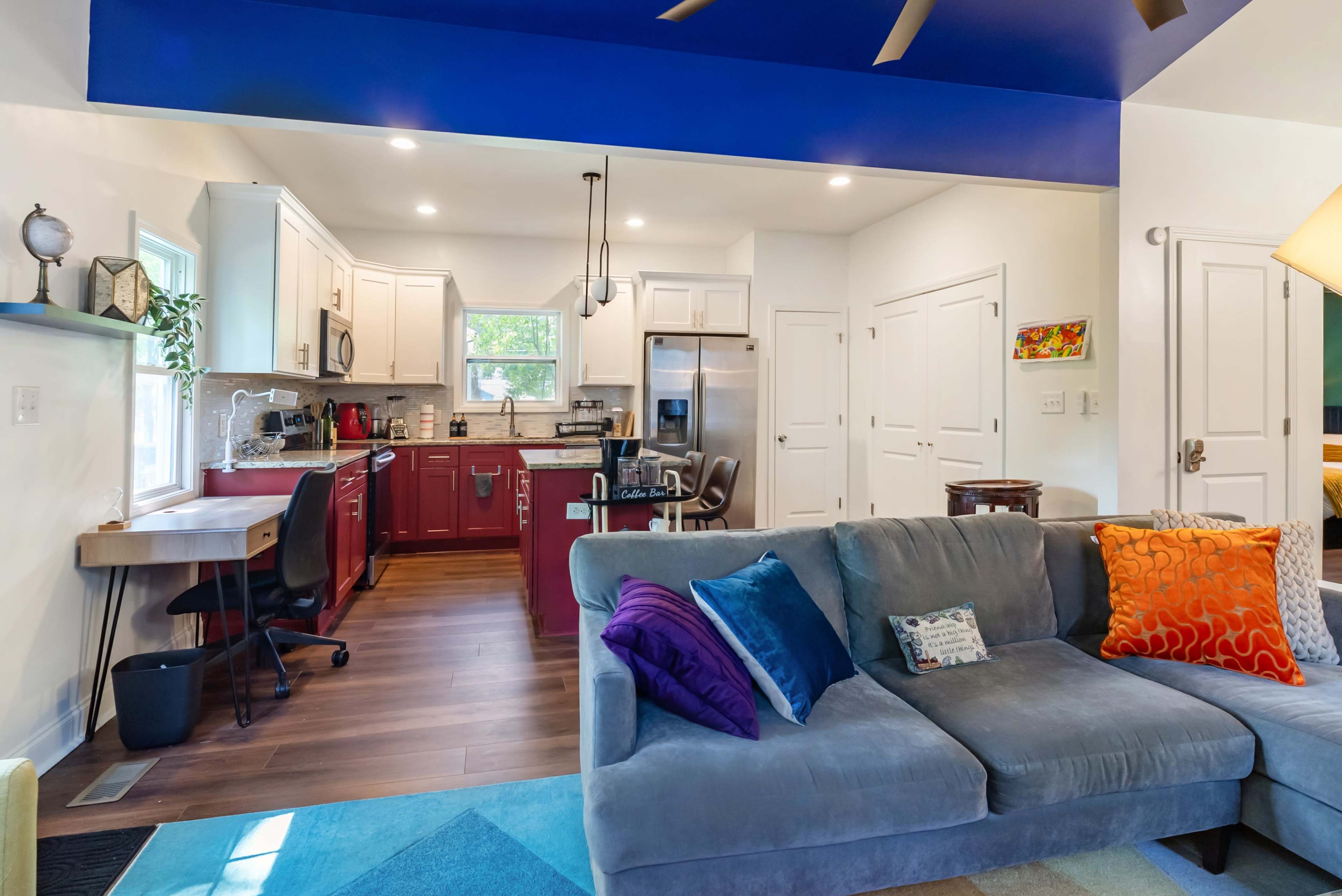 The image shows a modern living area with a gray sofa, colorful pillows, and an open kitchen featuring red cabinets and stainless steel appliances.