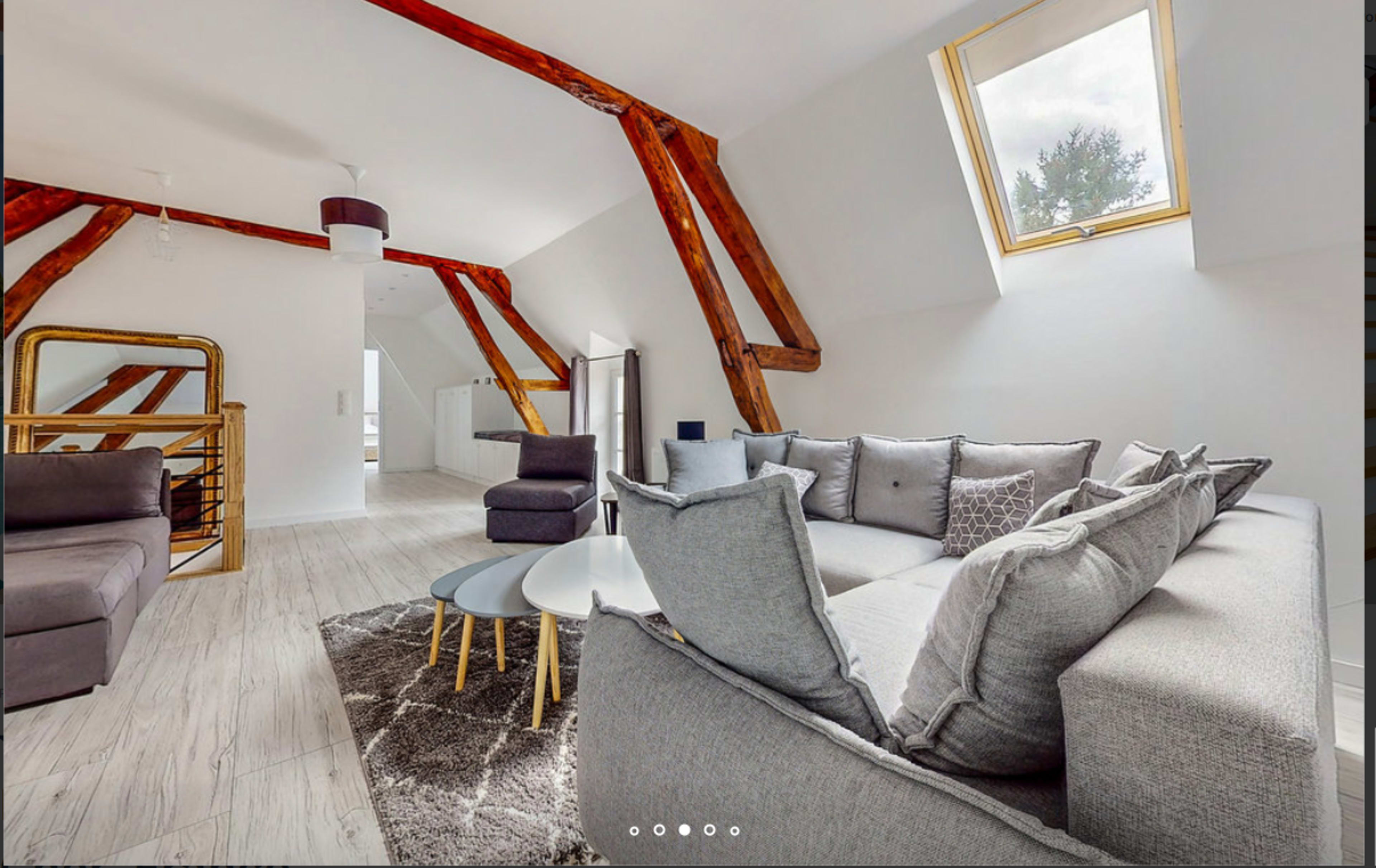 The image shows a modern living room featuring a gray sectional sofa, a round coffee table, and exposed wooden beams against a white wall.