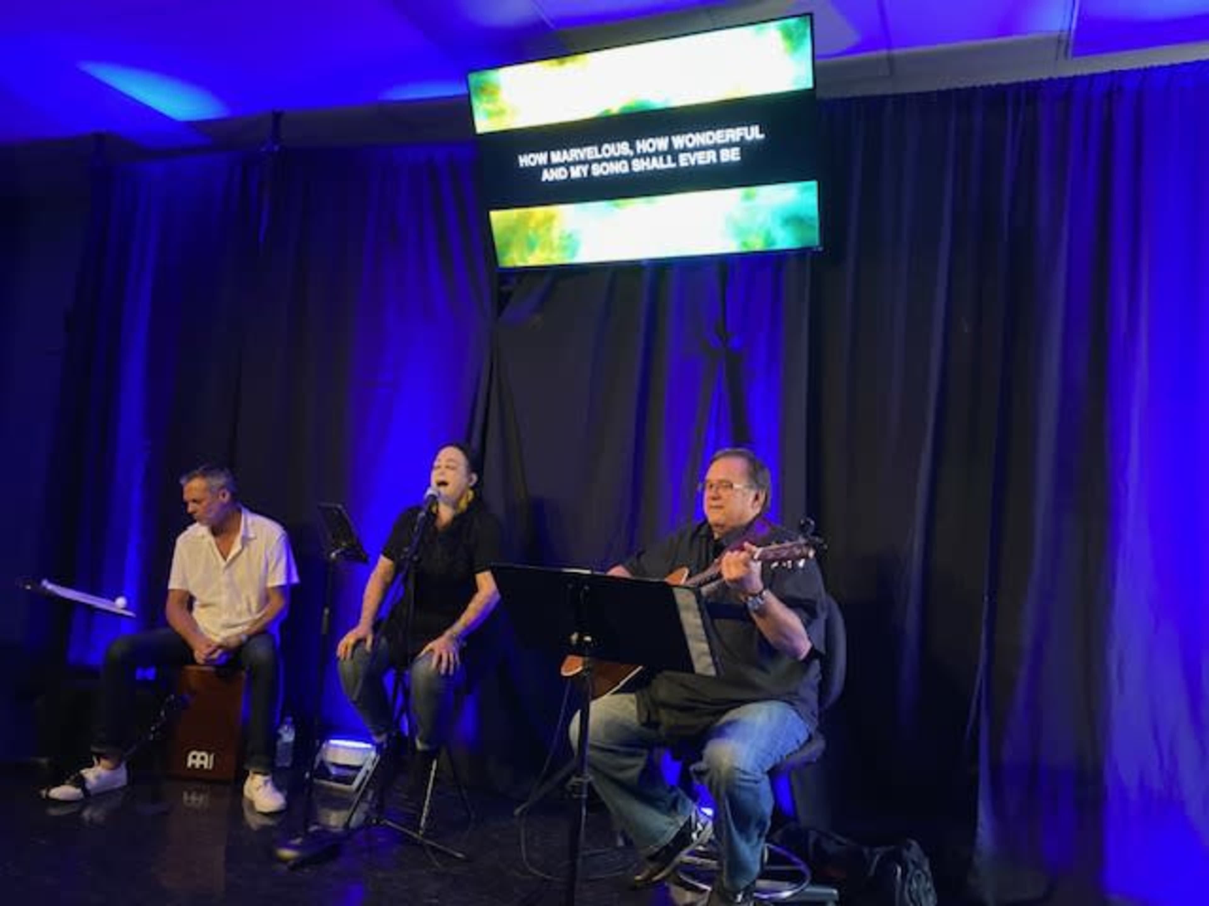 Three musicians perform on stage with a microphone and instruments, while song lyrics are displayed on a screen behind them.