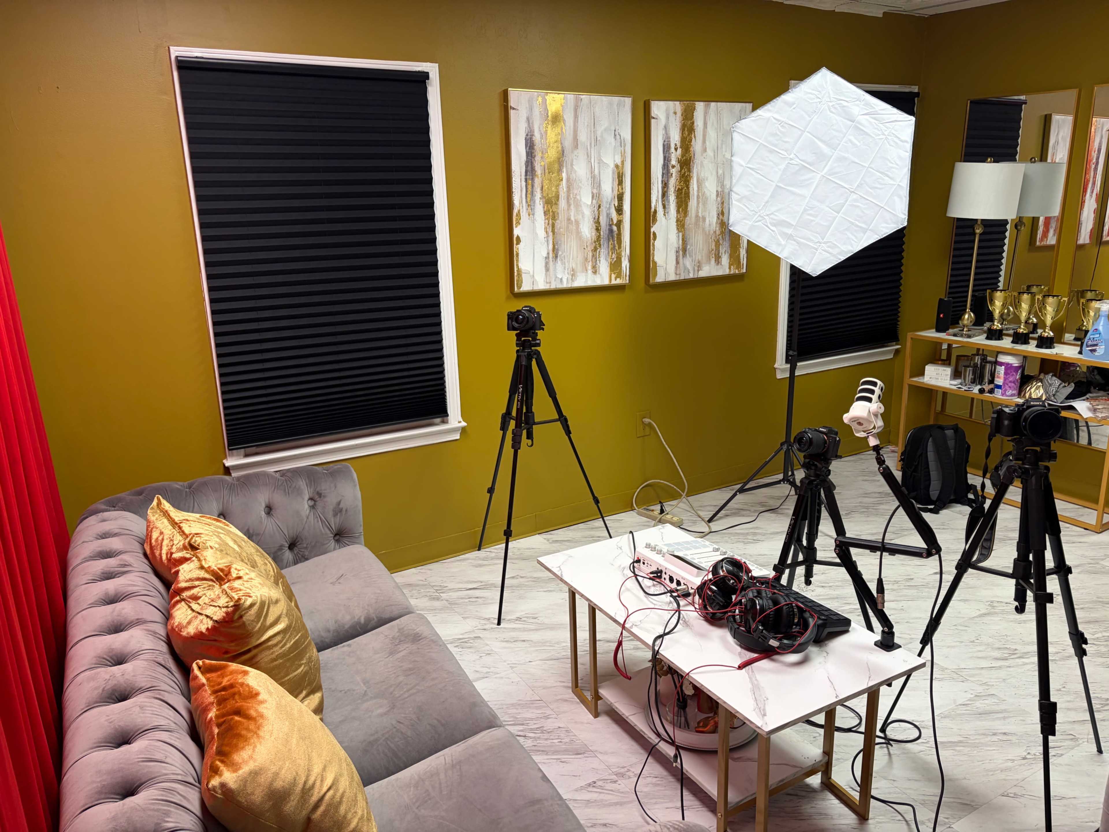 The image shows a room with a gray sofa, two black window blinds, a white table with tangled cables, and several tripods set up for photography or videography.