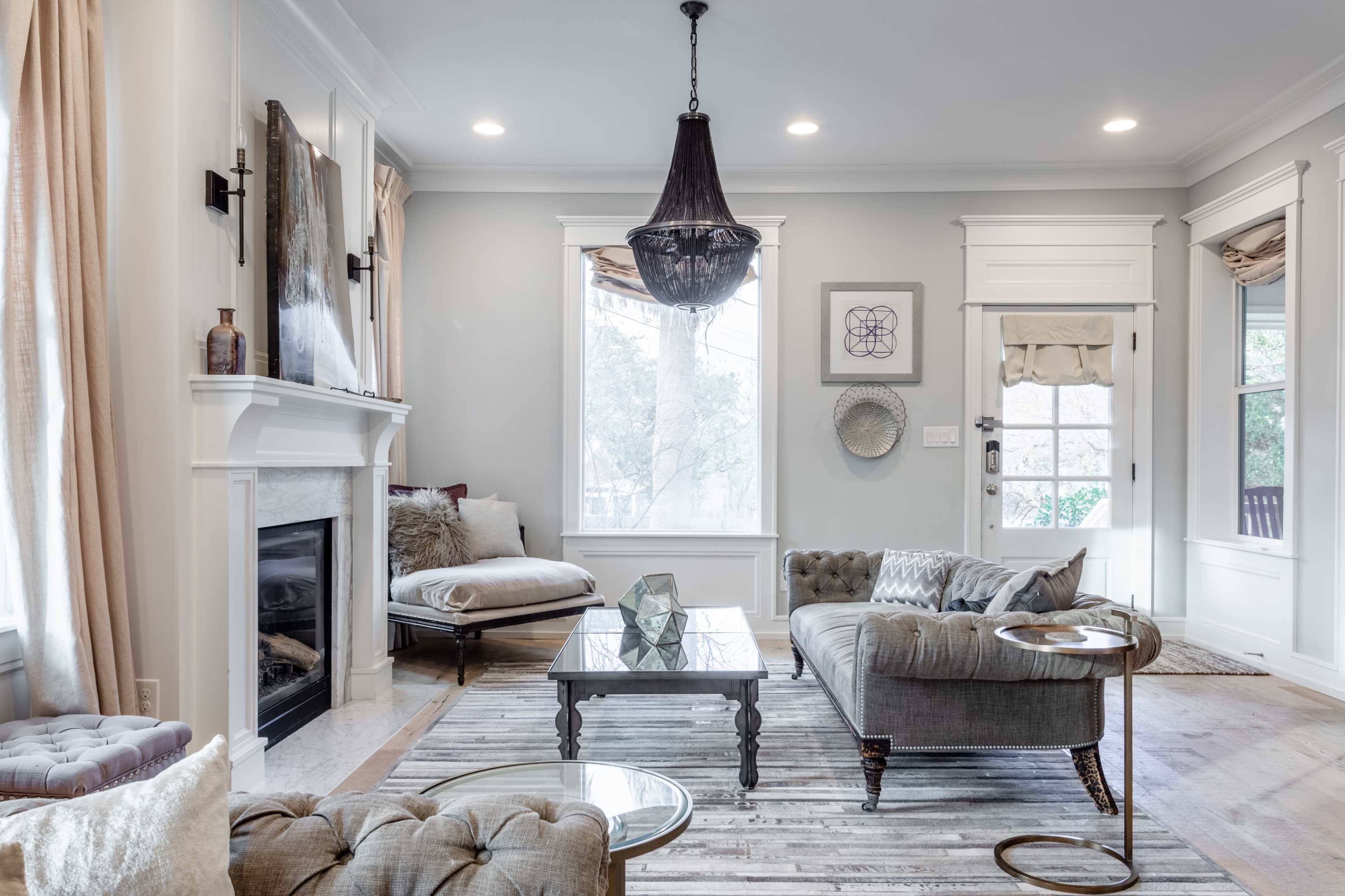 The living room features a light-colored design with a tufted gray sofa, a glass coffee table, and large windows allowing natural light to fill the space.