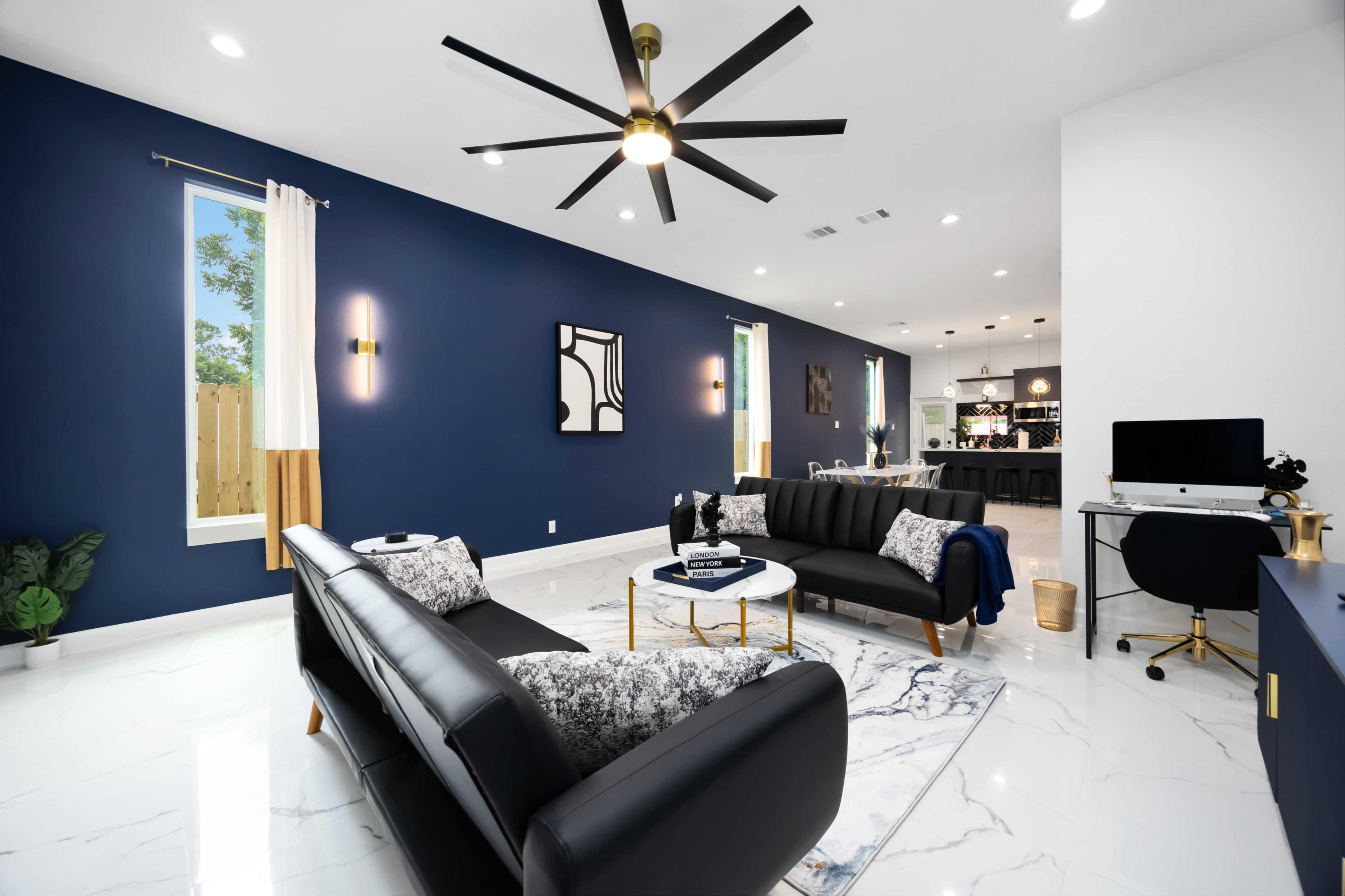 The living room features black sofas, a marble coffee table, and a large ceiling fan, with blue and white walls and a bright, open layout.