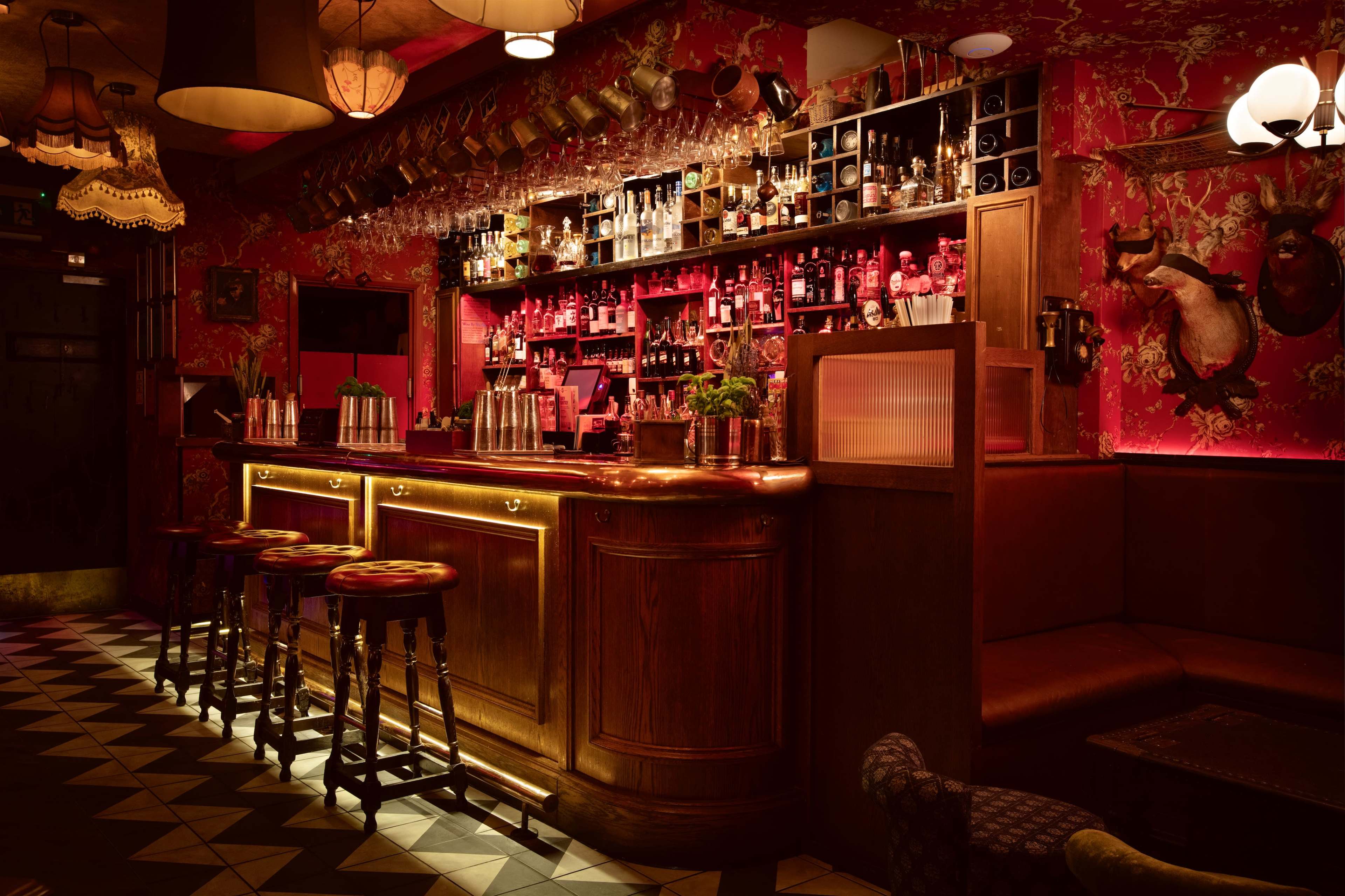 The image depicts a dimly lit bar with a wooden counter, surrounded by bar stools, and shelves lined with various bottles against a richly decorated, patterned wall.