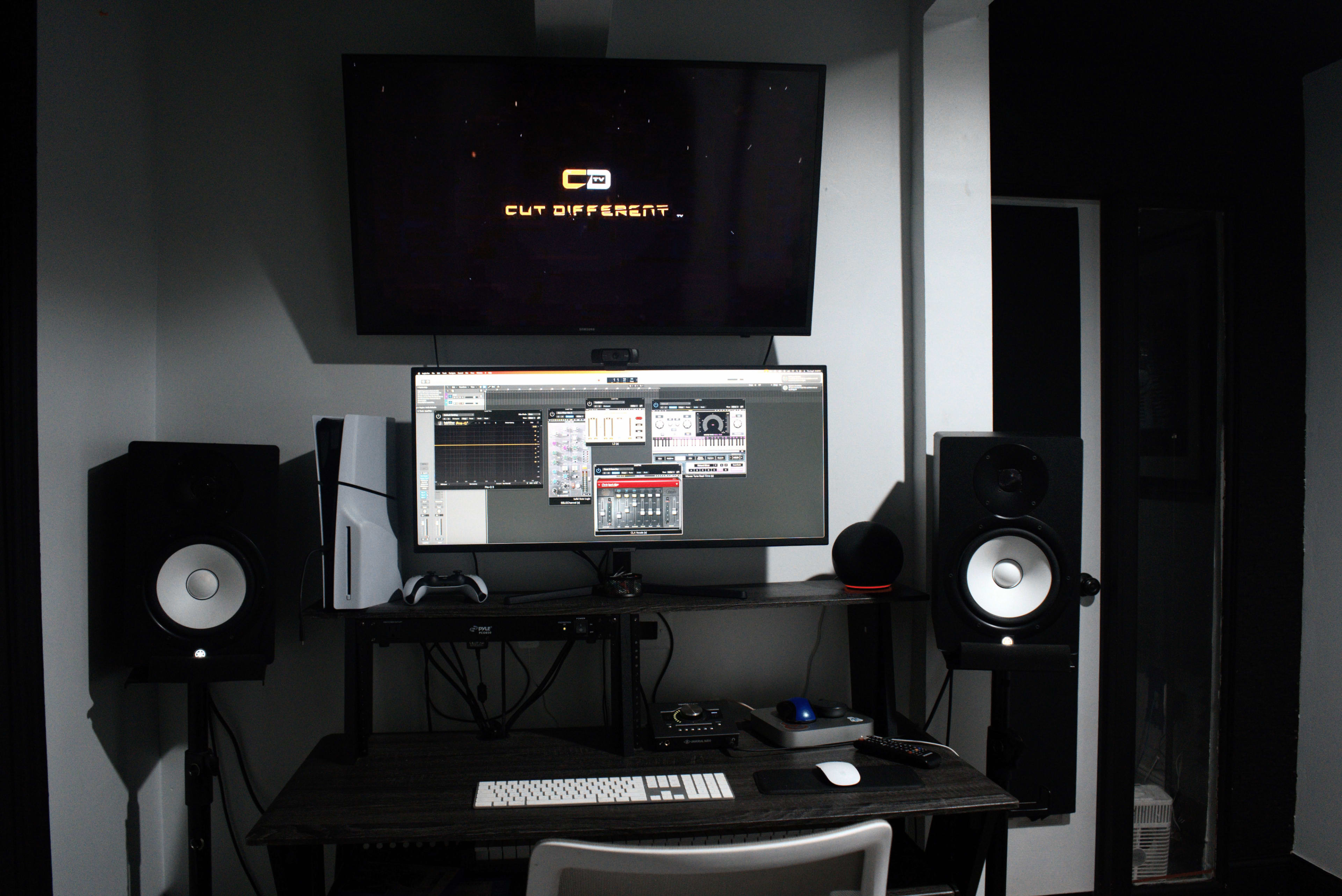 A computer workstation is set up with a large monitor displaying a music production software interface, flanked by studio speakers and a television mounted above.