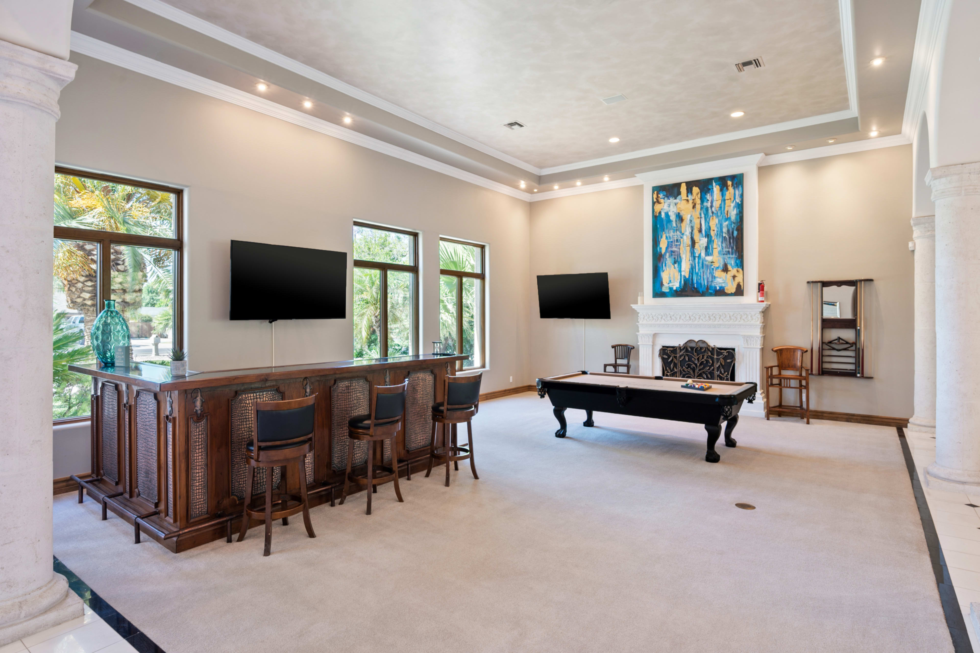 The image shows a spacious room with a bar area, a pool table, and large windows that provide natural light and a view of greenery outside.