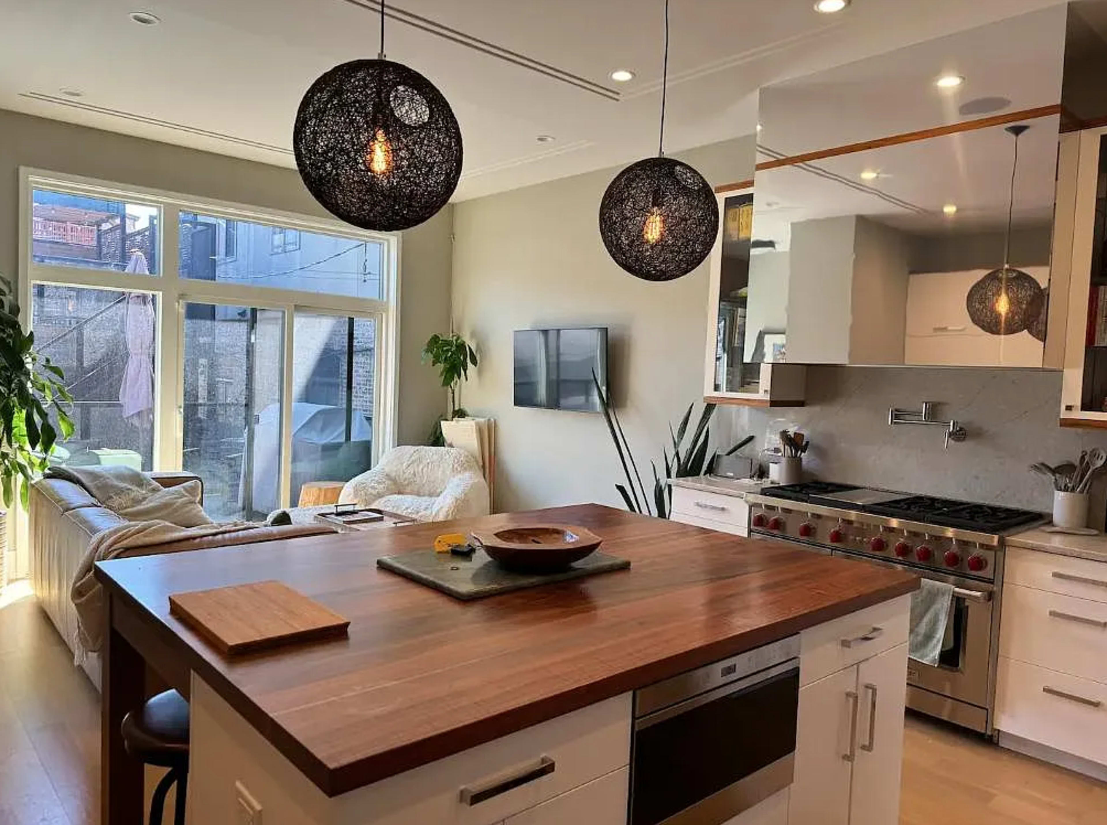 The image depicts a modern kitchen with an island featuring a wooden countertop, two pendant lights, and a cozy living area visible in the background.